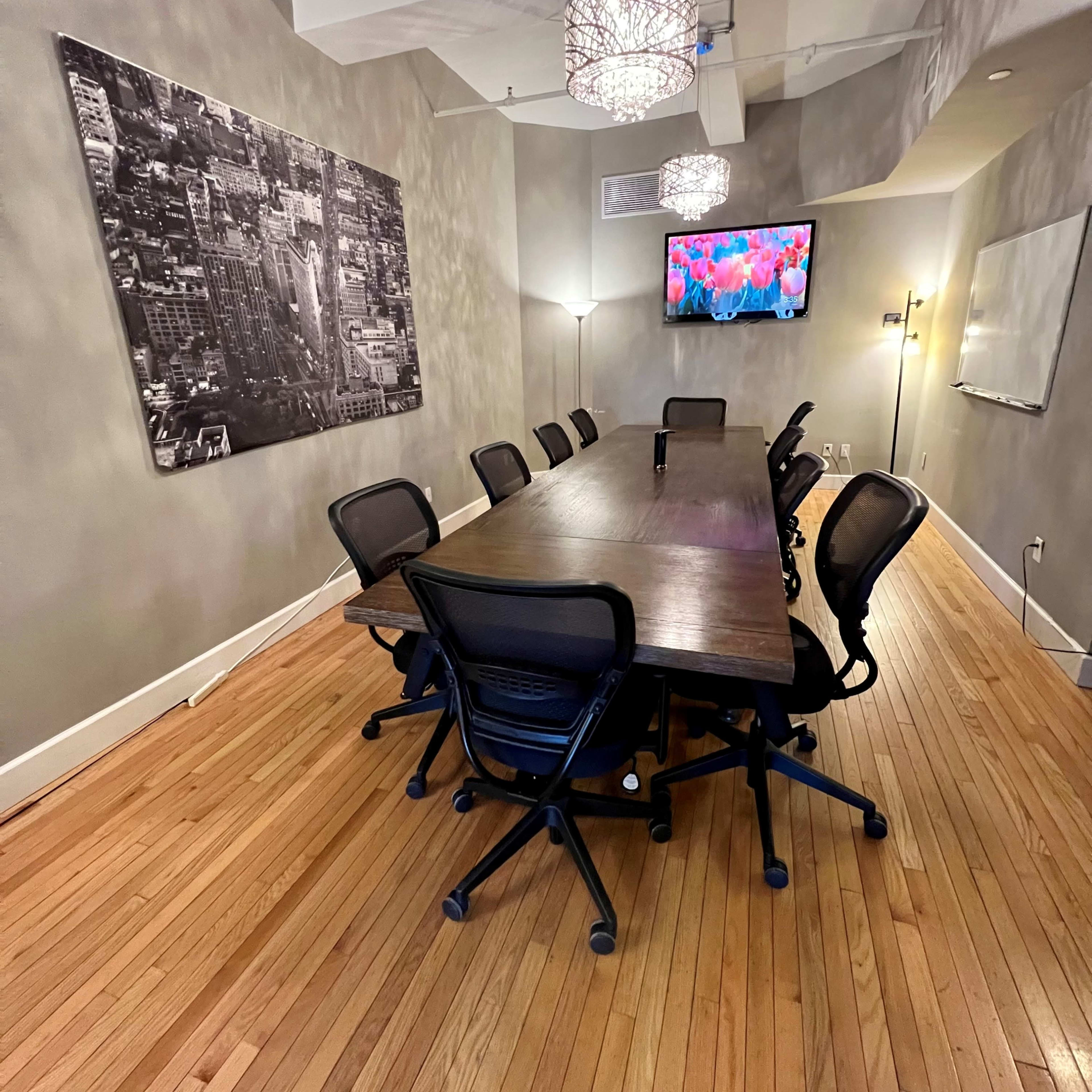 A long wooden conference table surrounded by ergonomic chairs is set in a well-lit meeting room featuring a large wall photo and a screen displaying vibrant images.