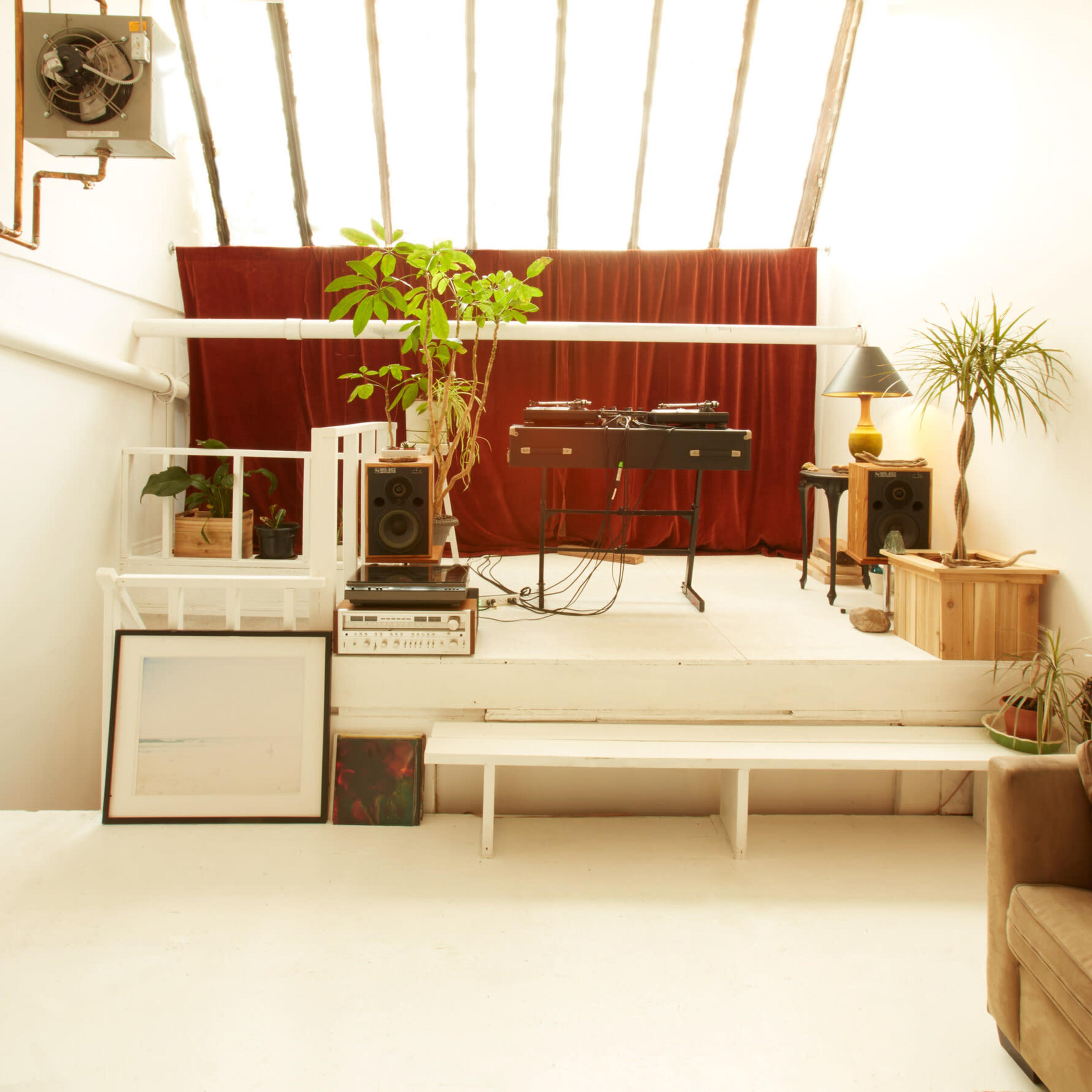 A bright, minimalistic interior with a raised platform featuring a DJ setup, plants, and various pieces of furniture against a backdrop of red curtains.