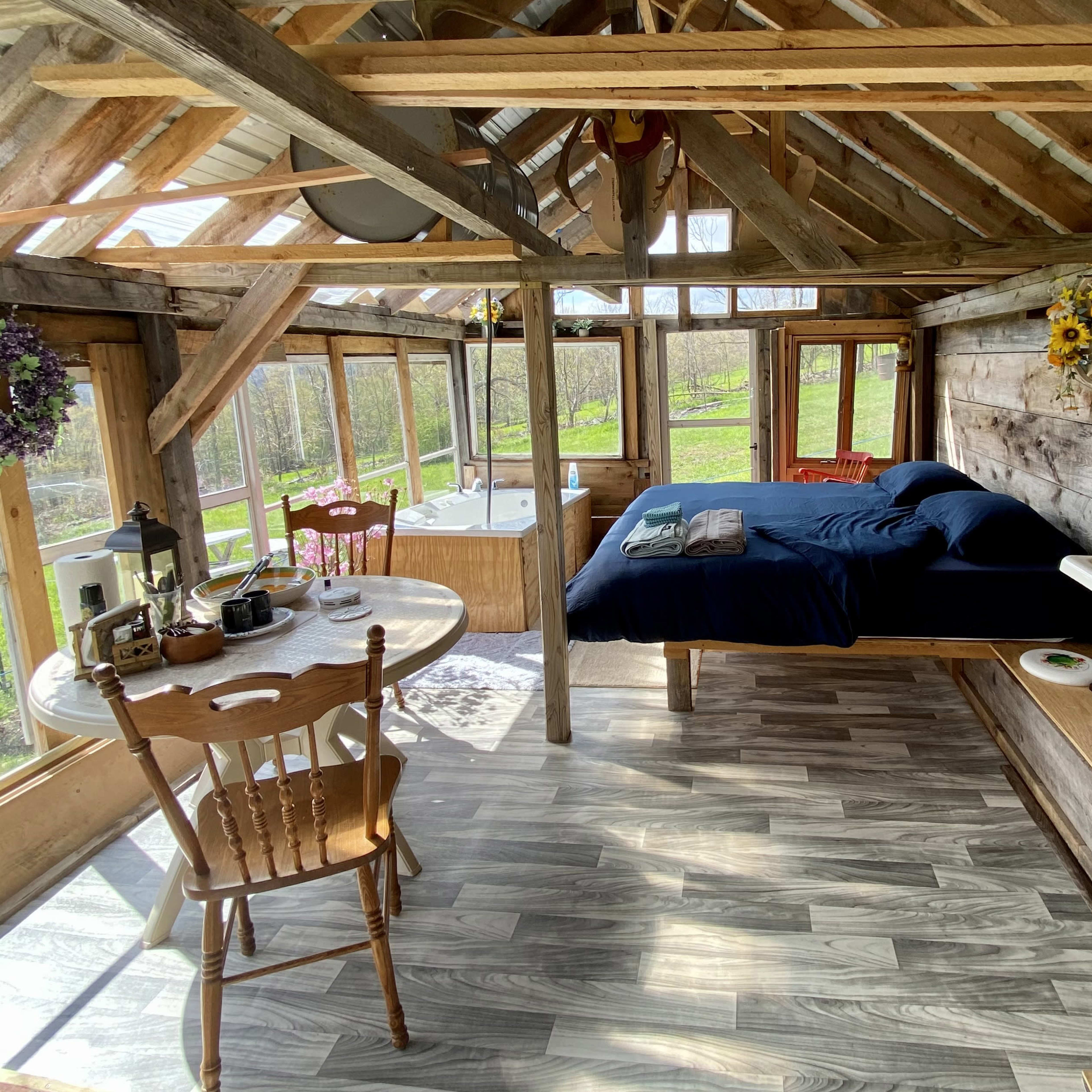 A rustic wooden cabin interior featuring a bed, a dining table with chairs, and a large window area overlooking a grassy landscape.