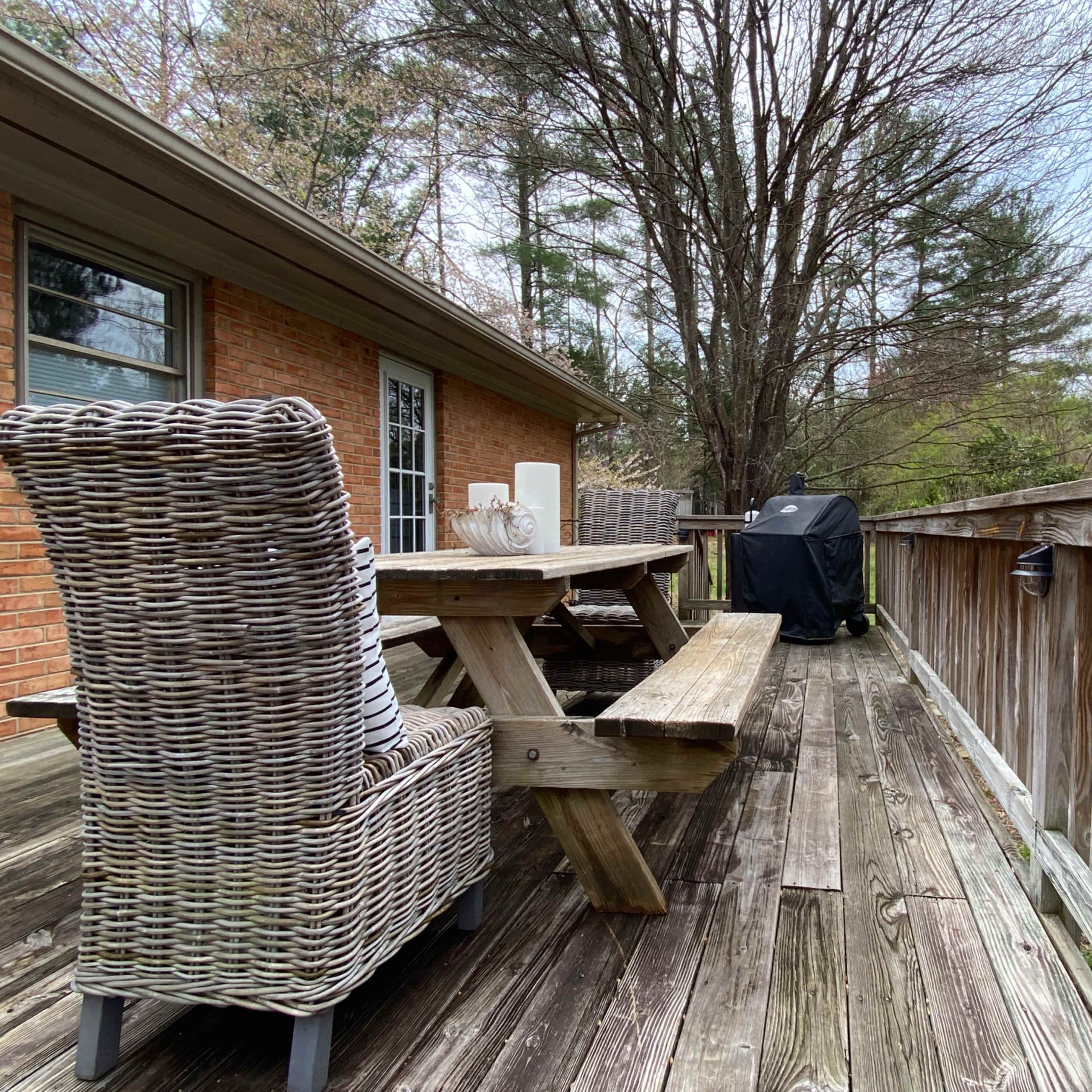 A wooden deck features a picnic table, a wicker chair, and a grill surrounded by trees.