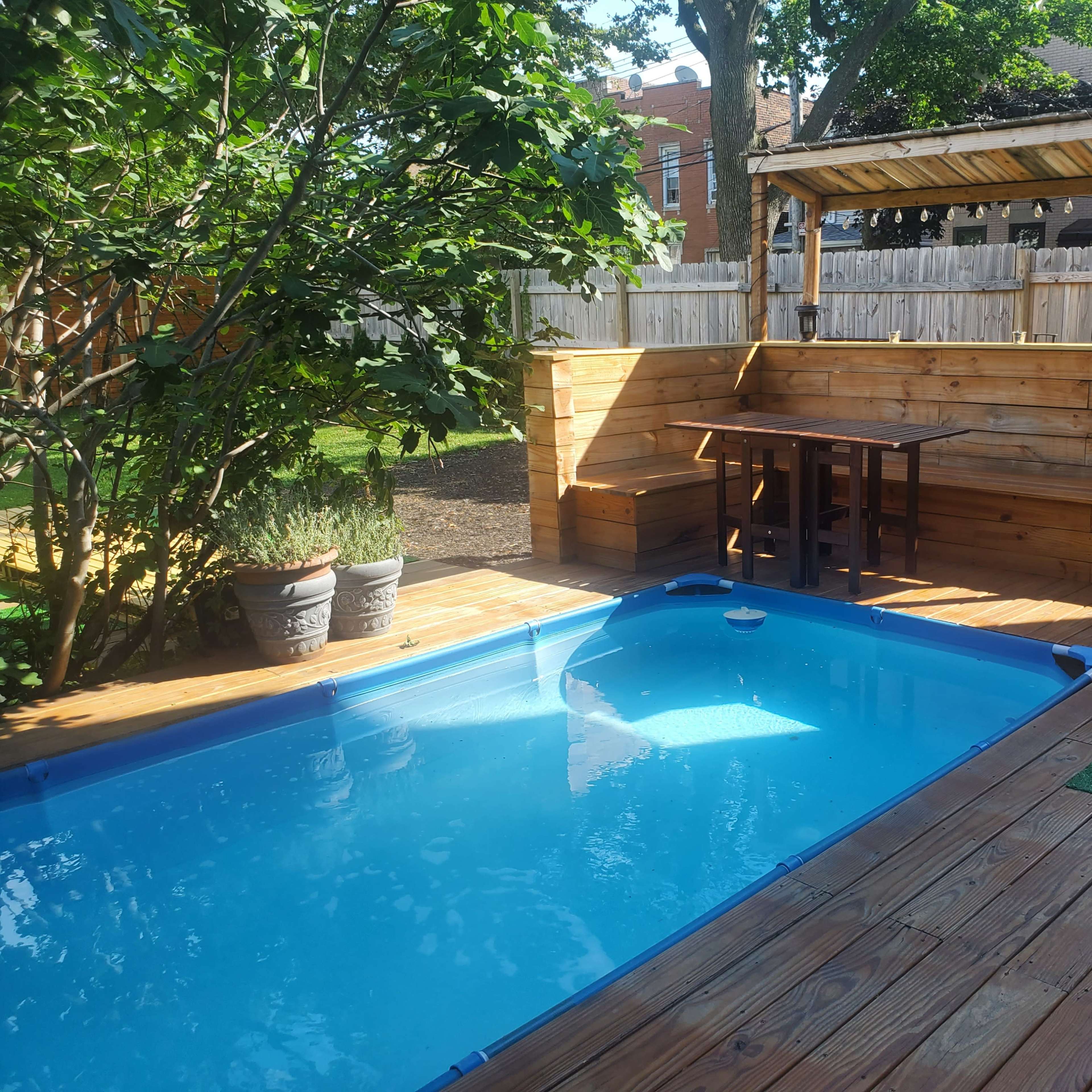 A small backyard featuring a blue pool, a wooden seating area with a table, and green plants within a fenced enclosure.