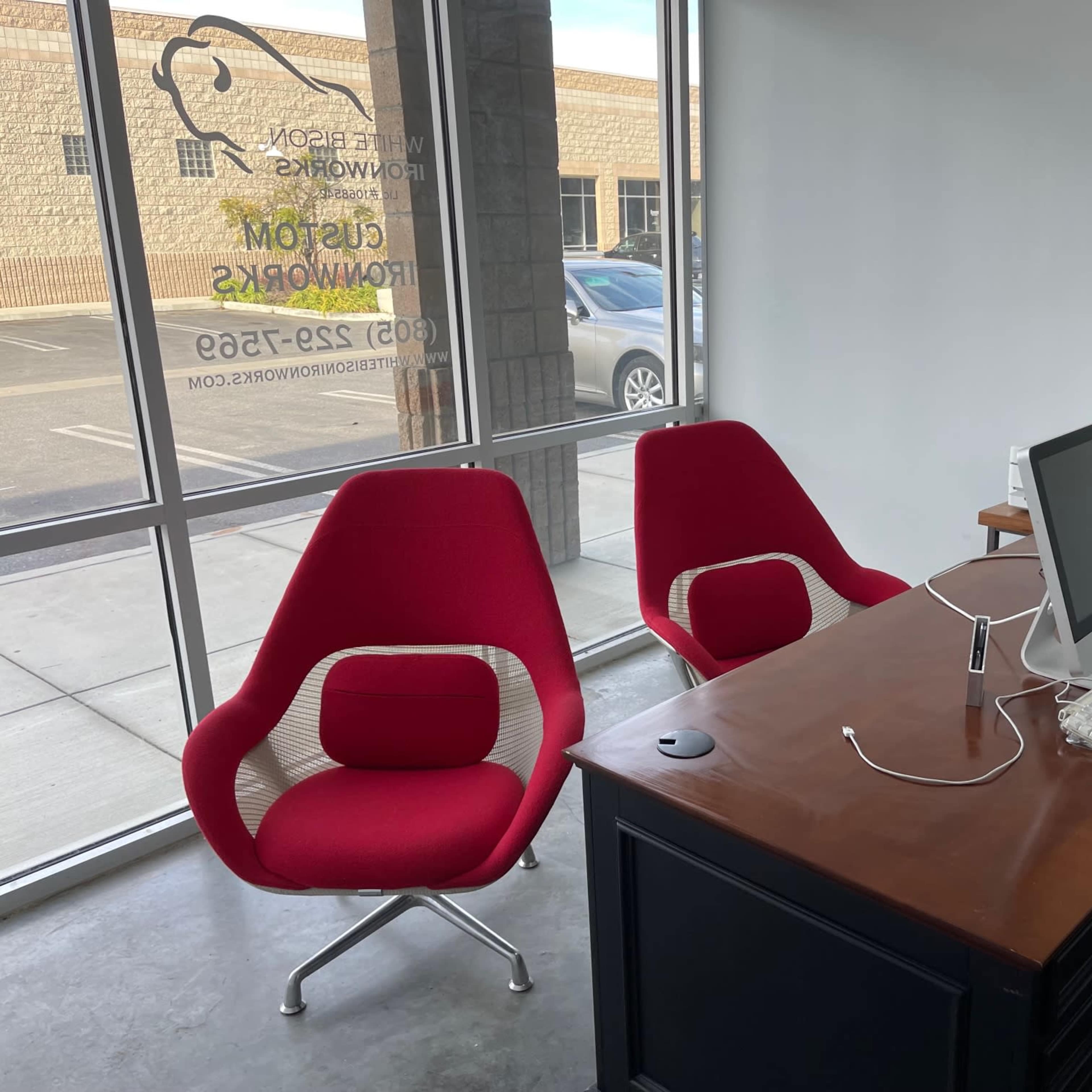 A modern office space with two red chairs facing a wooden desk that holds a computer and various office supplies, set against a large window.