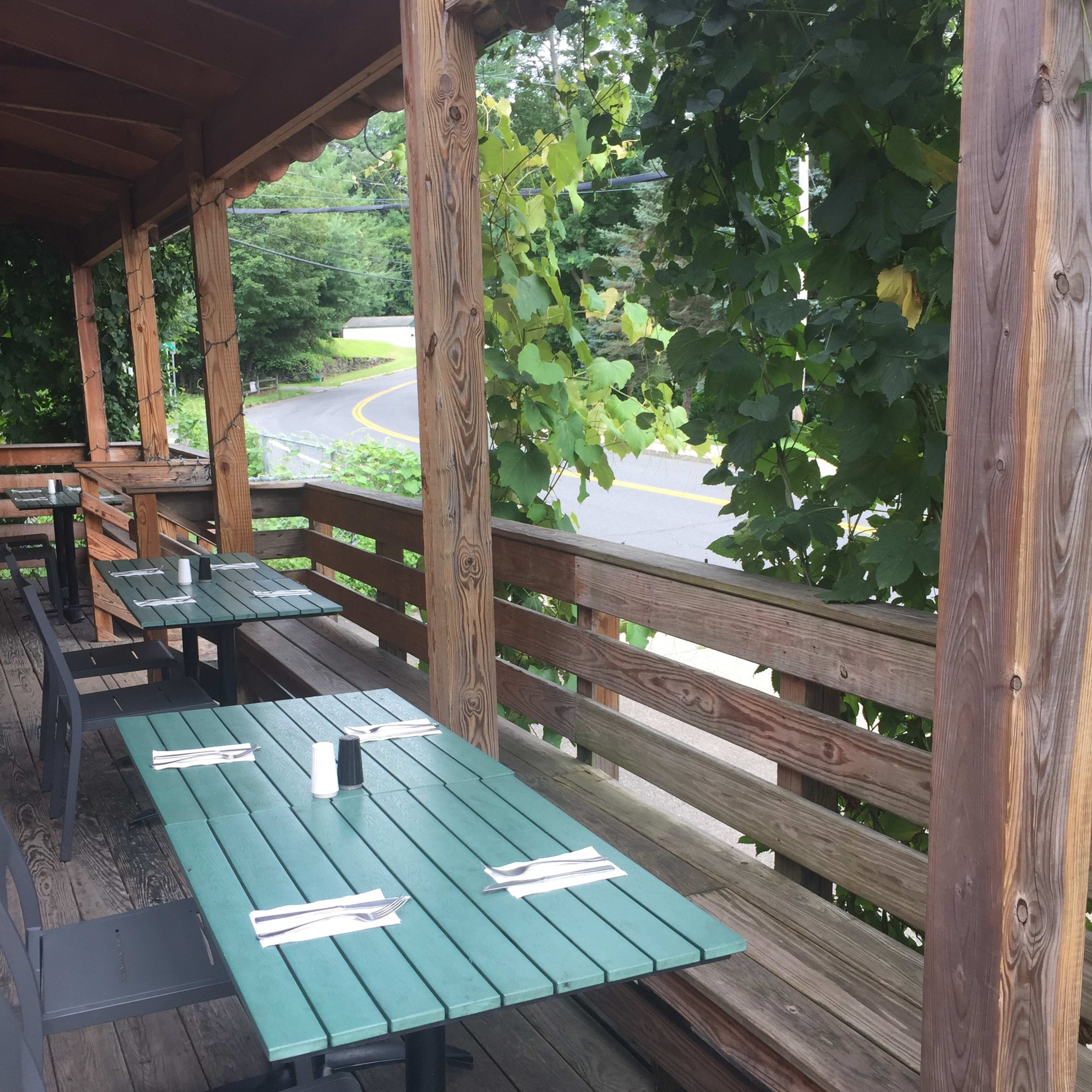 A patio area of a restaurant with wooden tables set for dining under a wooden awning, surrounded by greenery.