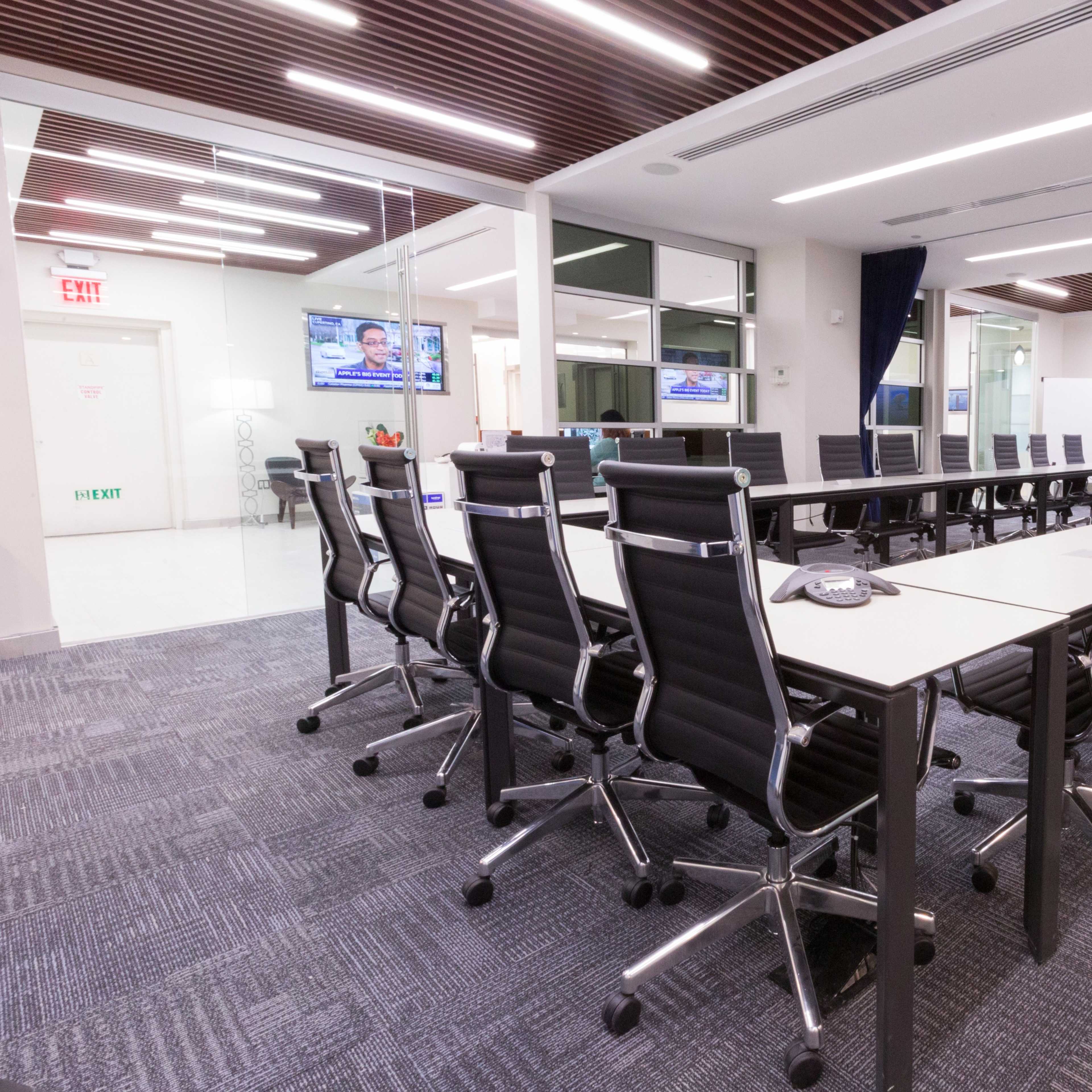 A long conference table surrounded by black office chairs is positioned in a well-lit meeting room featuring glass walls and multiple screens.