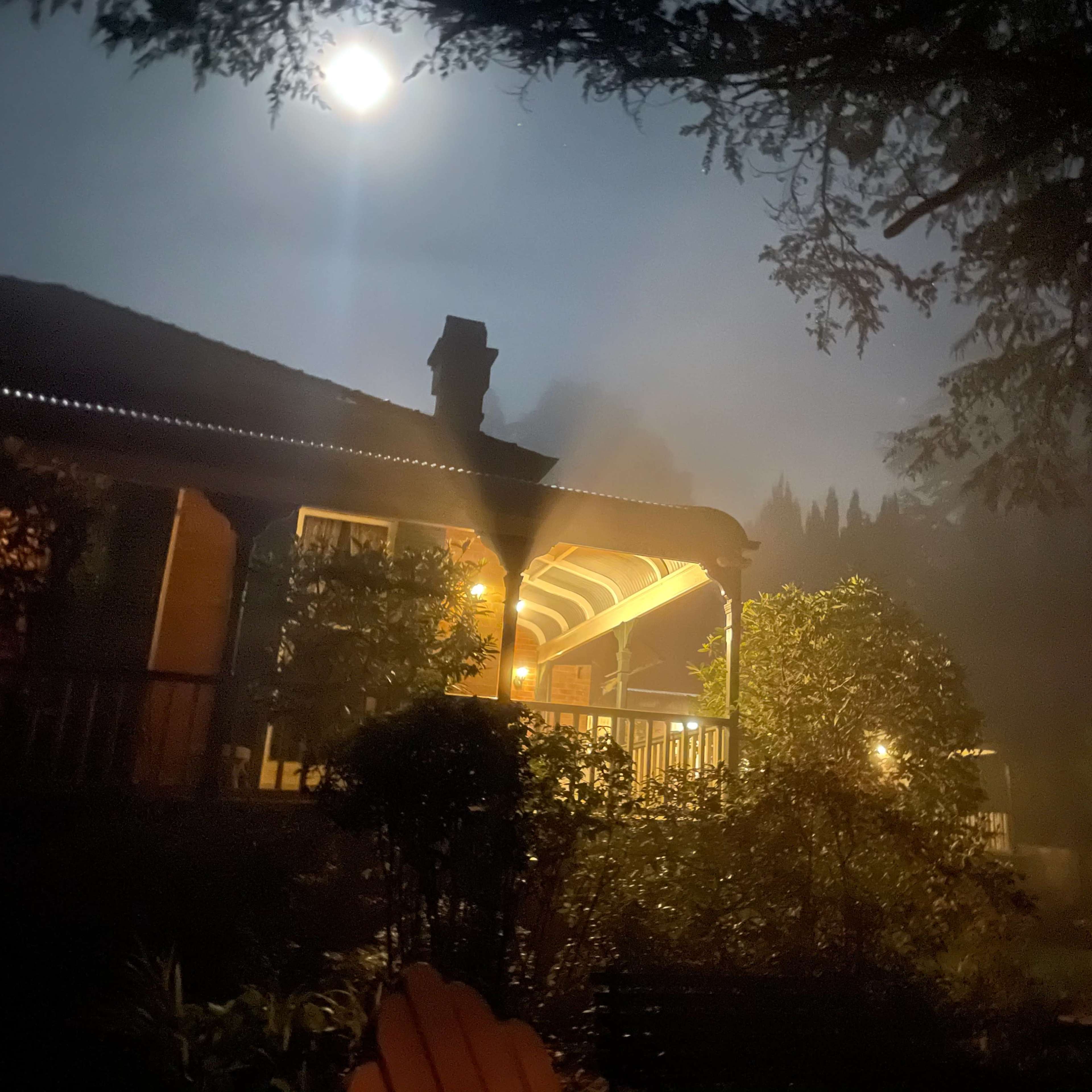 A well-lit porch stands in the foreground under a full moon, surrounded by fog and trees.