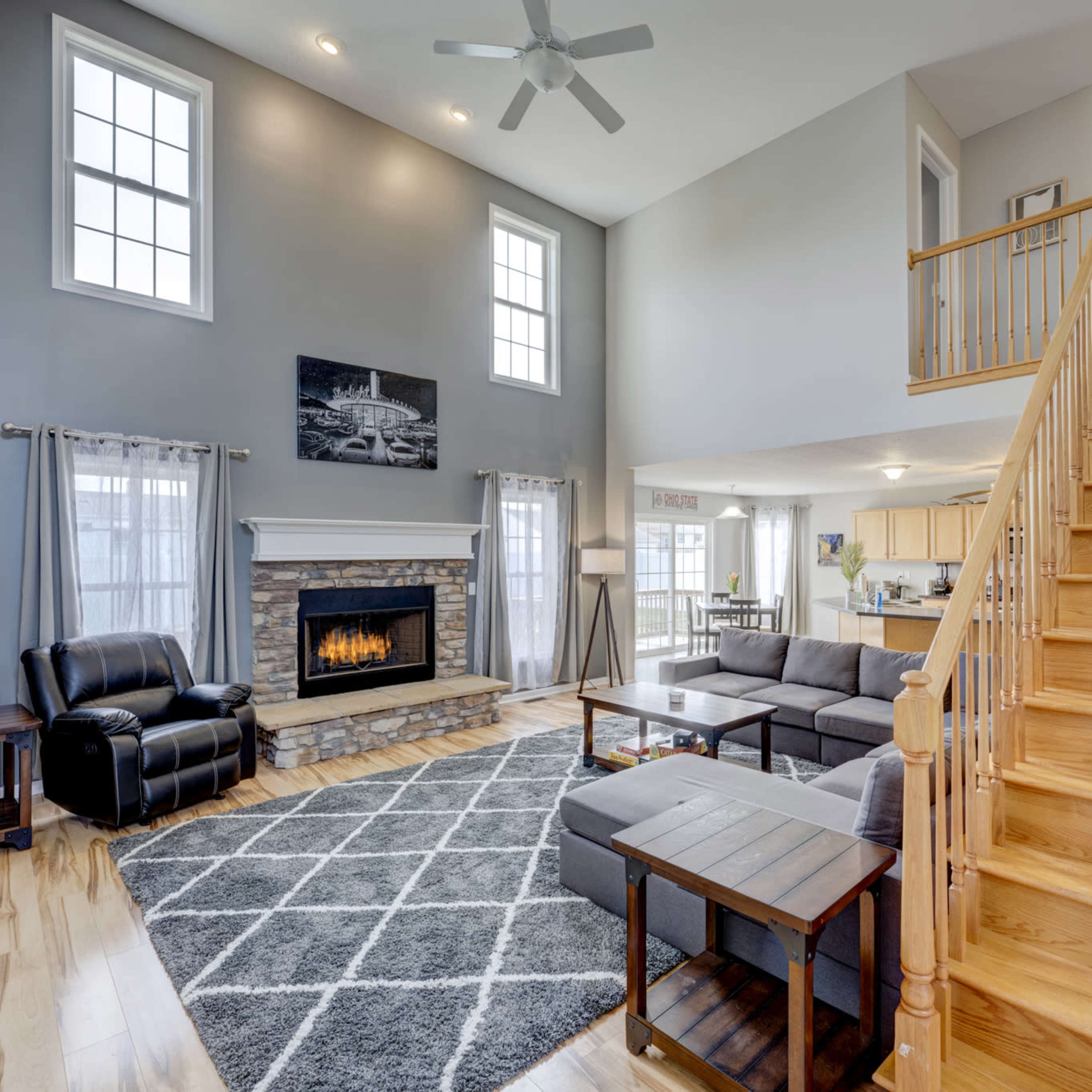 A two-story living room with a fireplace, large windows, a gray sectional sofa, and a wooden staircase leading to the upper level.