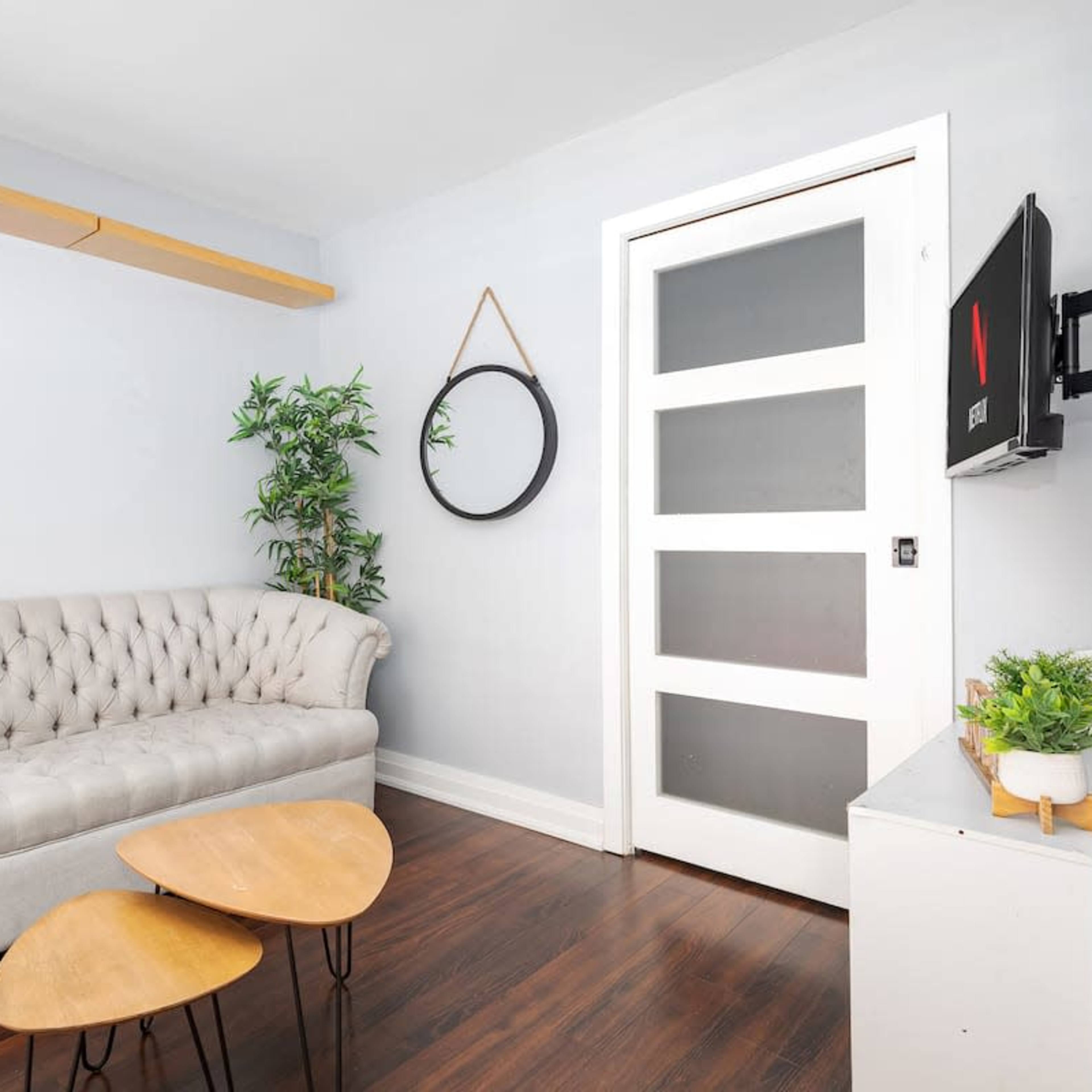 A light-colored room features a tufted gray sofa, a round mirror on the wall, a small plant, and a wooden coffee table.
