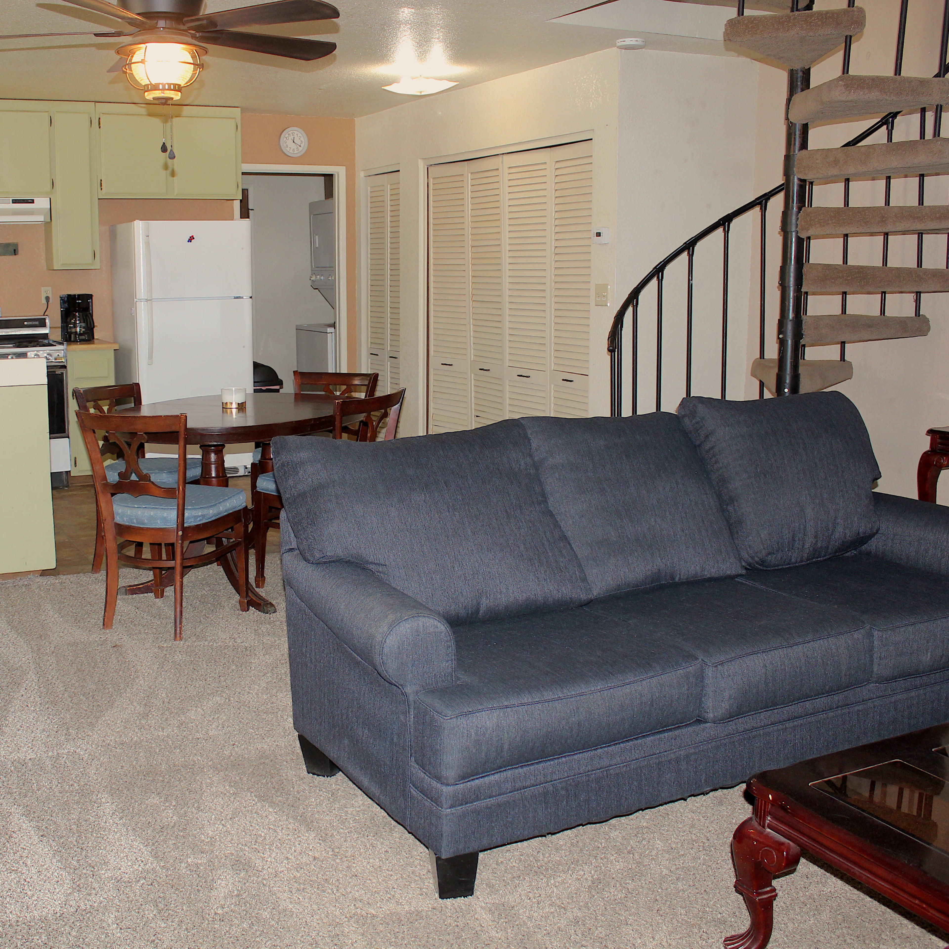 A living area with a blue couch, a dining table with chairs, a kitchen in the background, and a spiral staircase.