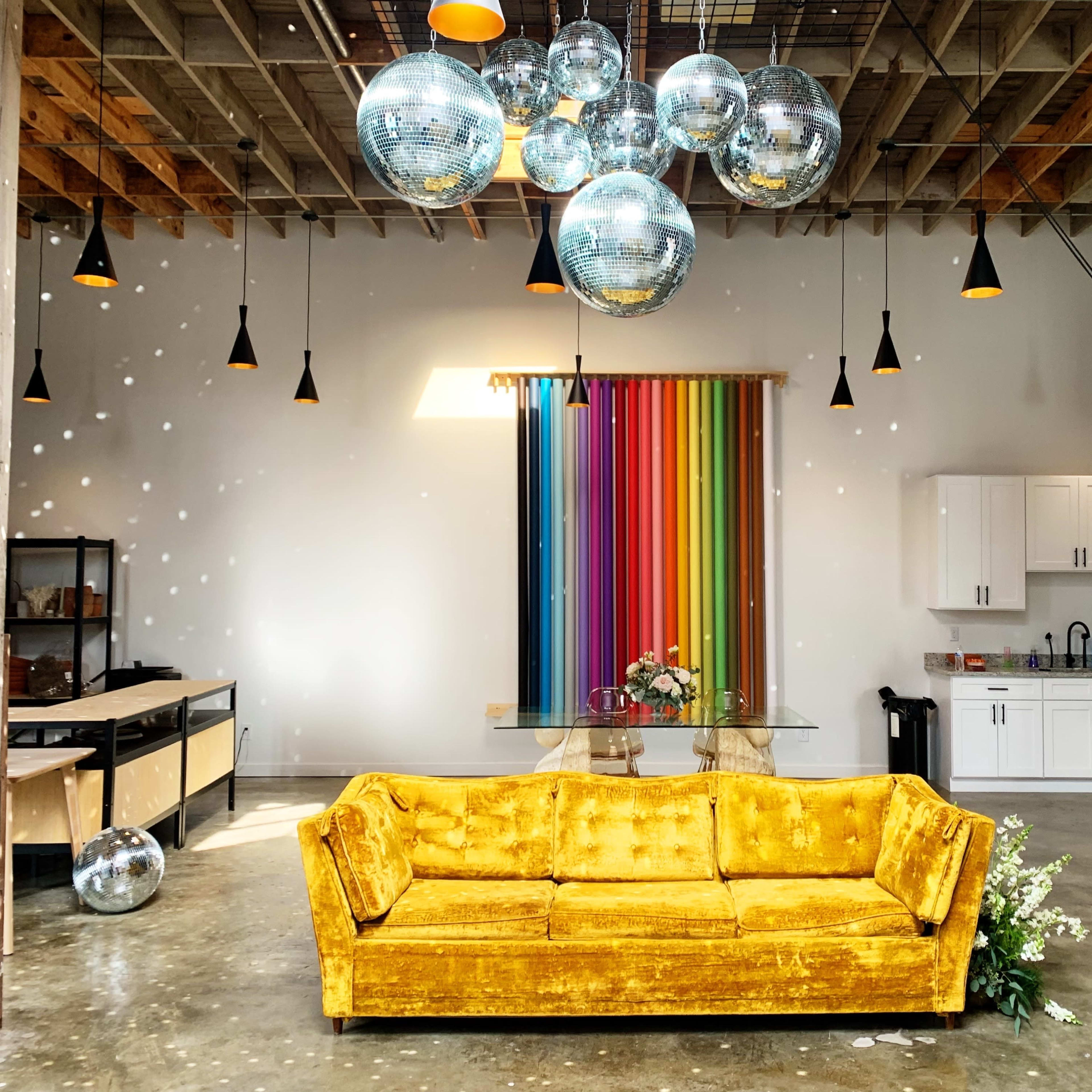 A yellow velvet couch is centered in a spacious room with a colorful striped backdrop, disco balls hanging from the ceiling, and a modern kitchen in the background.