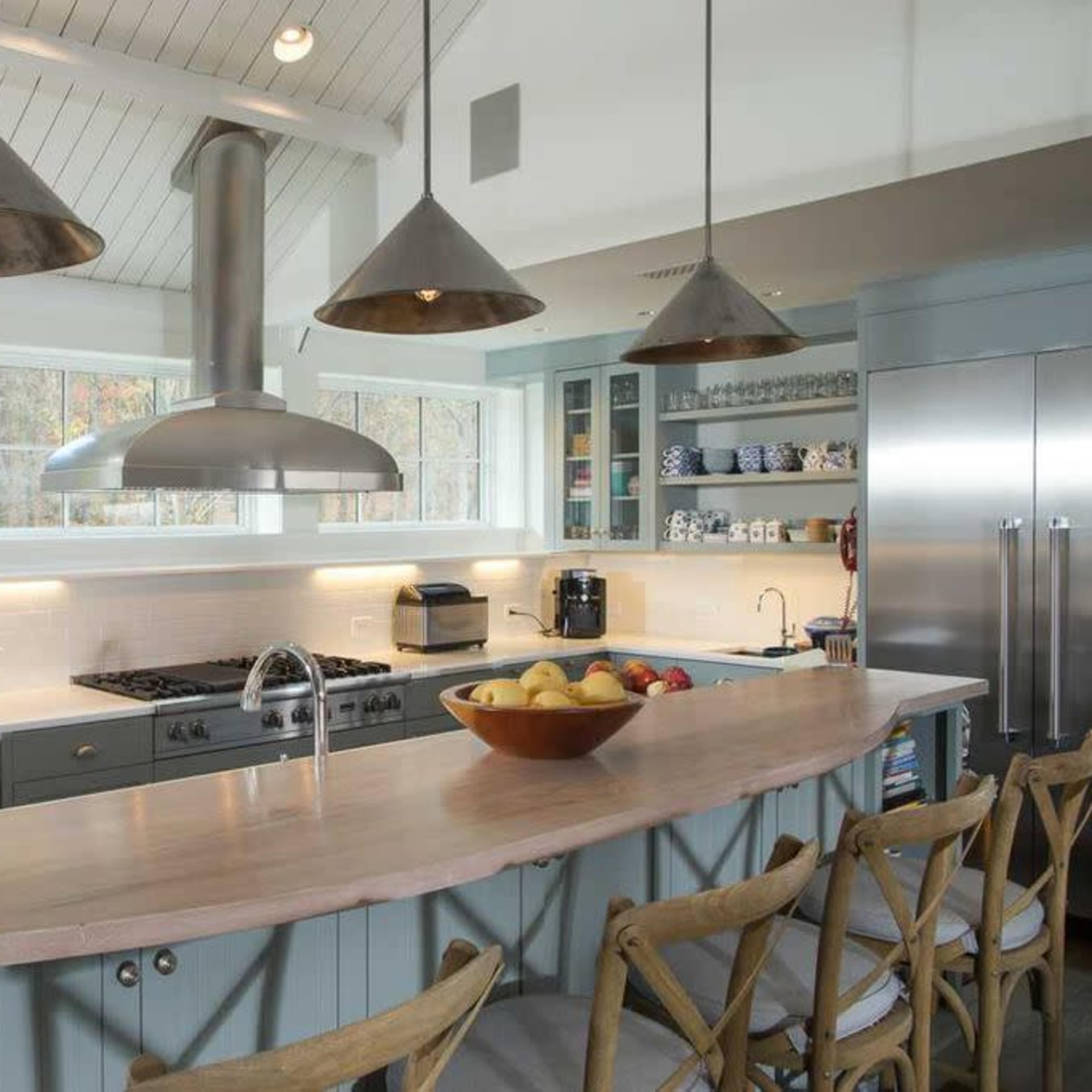 A modern kitchen features a central island with a curved wooden countertop, three pendant lights overhead, and stainless steel appliances along one wall.