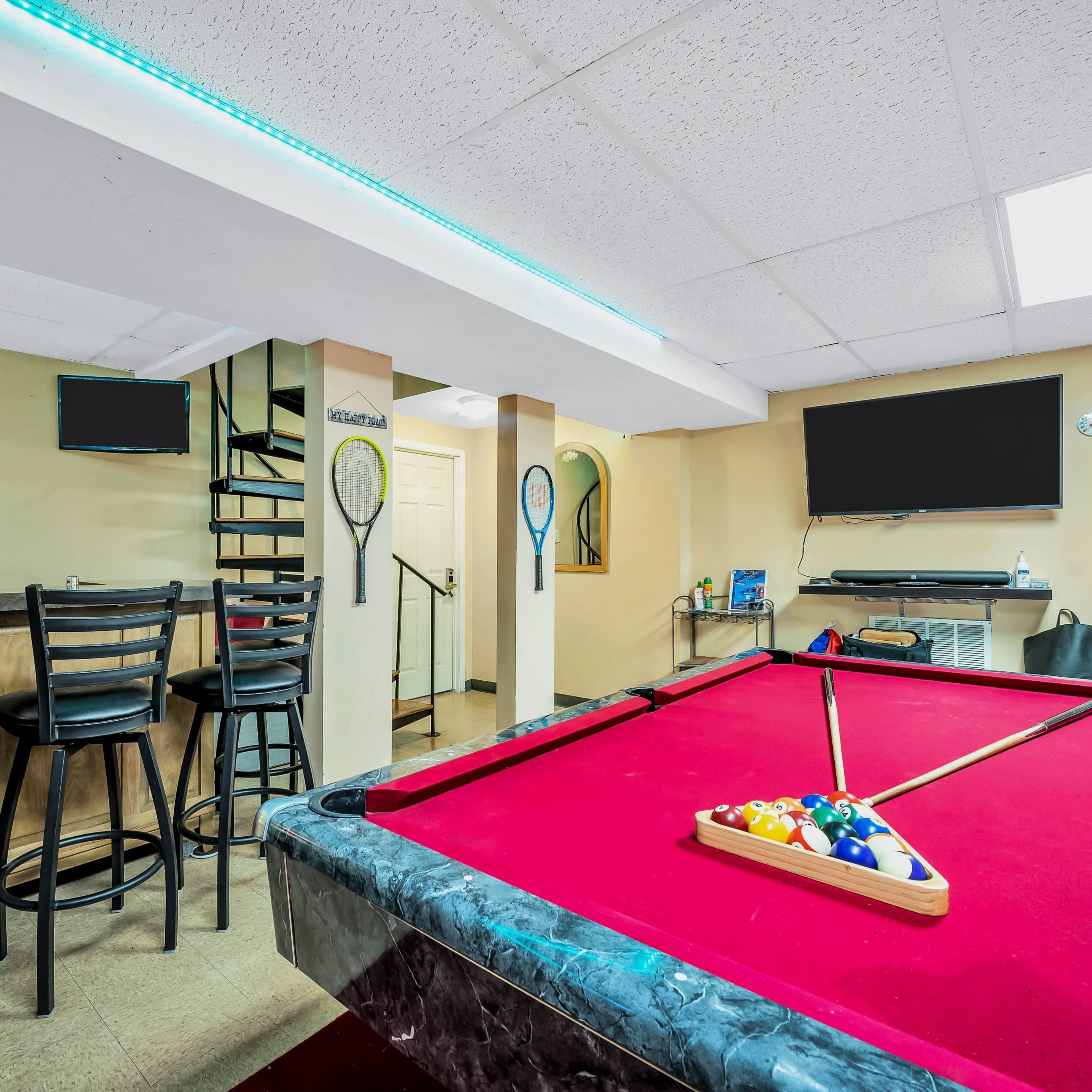 A basement game room featuring a red pool table, bar stools, a television, and several staircase accesses.