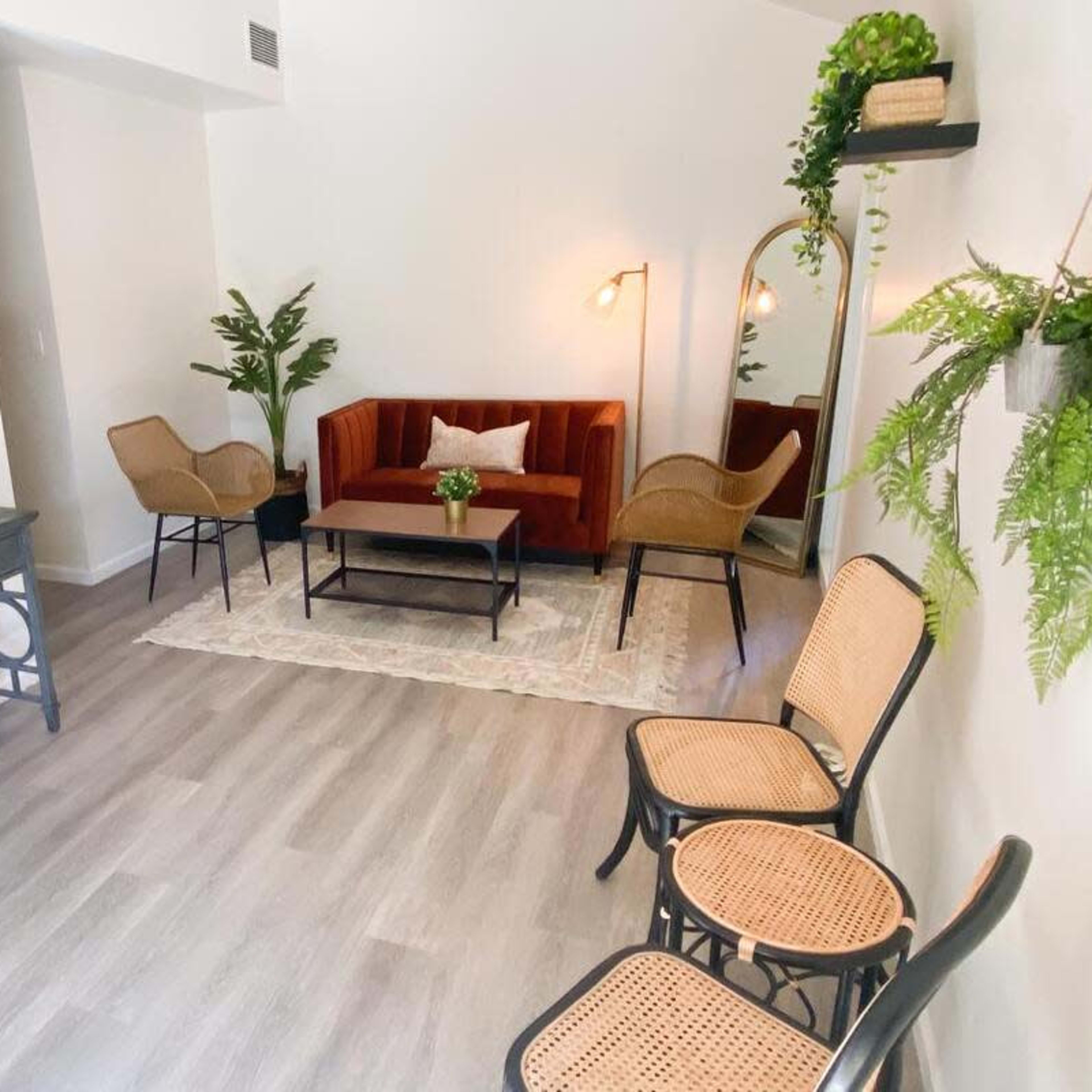 The room features a red velvet sofa, two cane chairs, a coffee table, and a floor mirror, with plants and decorative items arranged throughout.