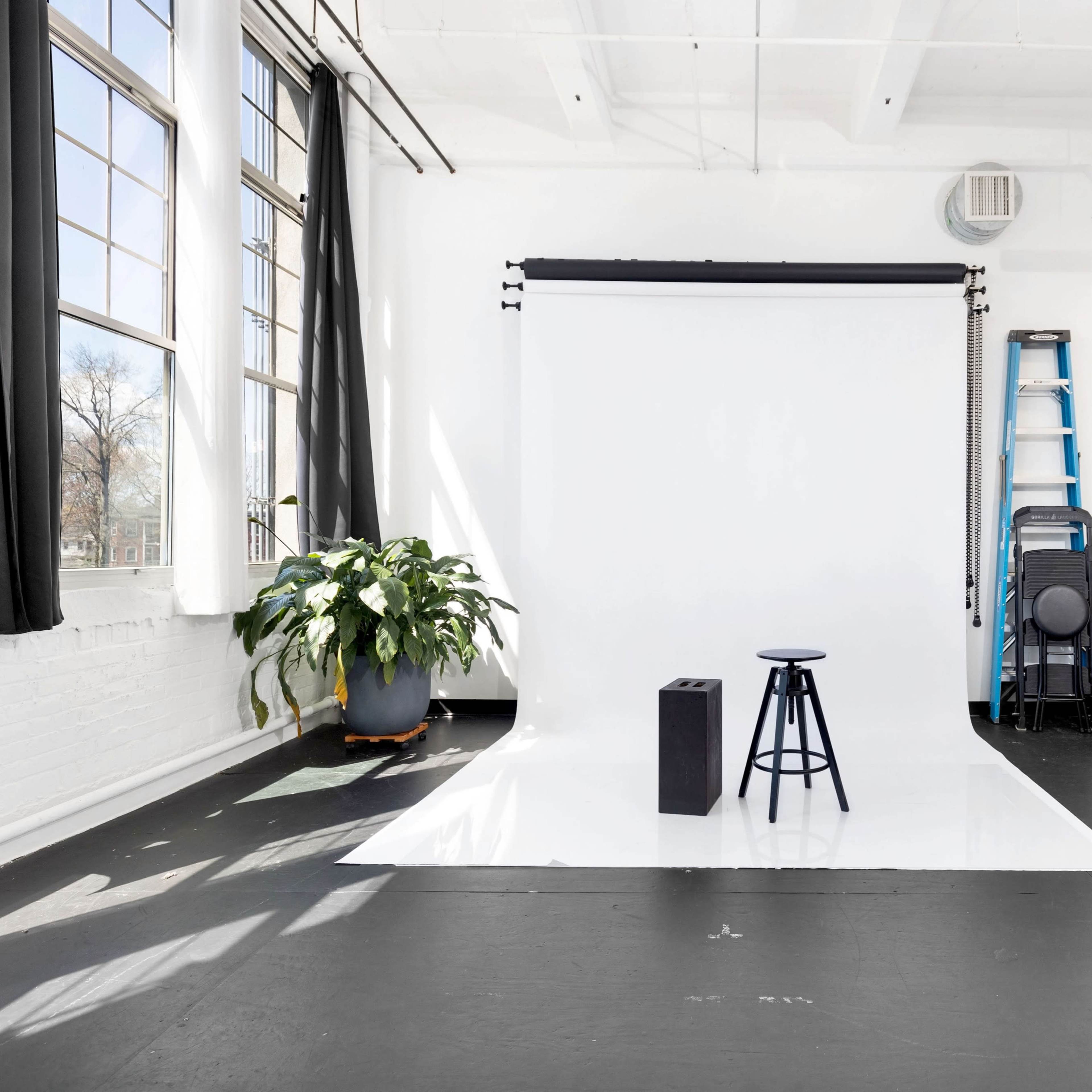 A photography studio featuring a white backdrop, a stool, a black box, and plants along with large windows and a ladder.