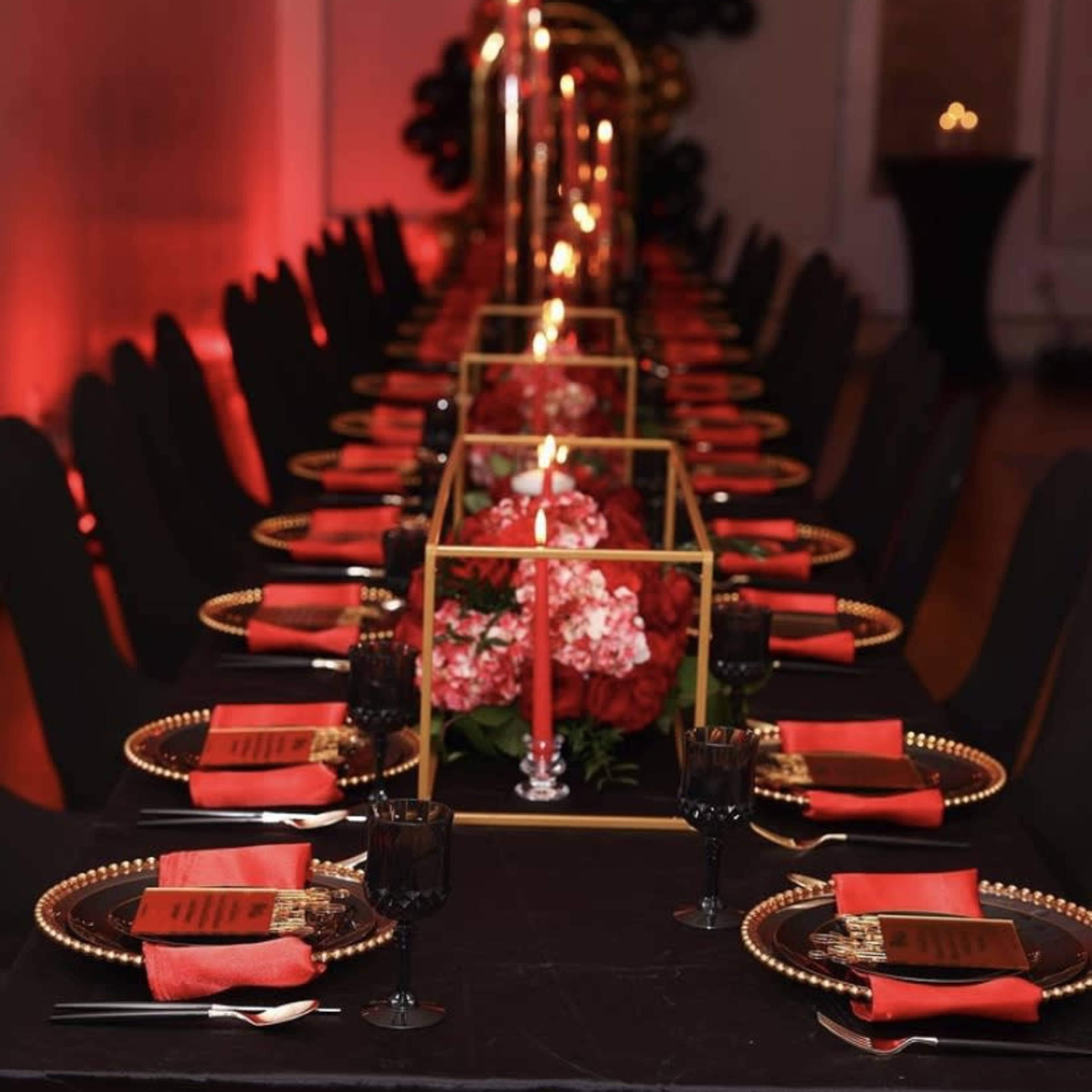 A long dining table is elegantly set with black and gold plates, red napkins, and floral centerpieces, illuminated by candles.