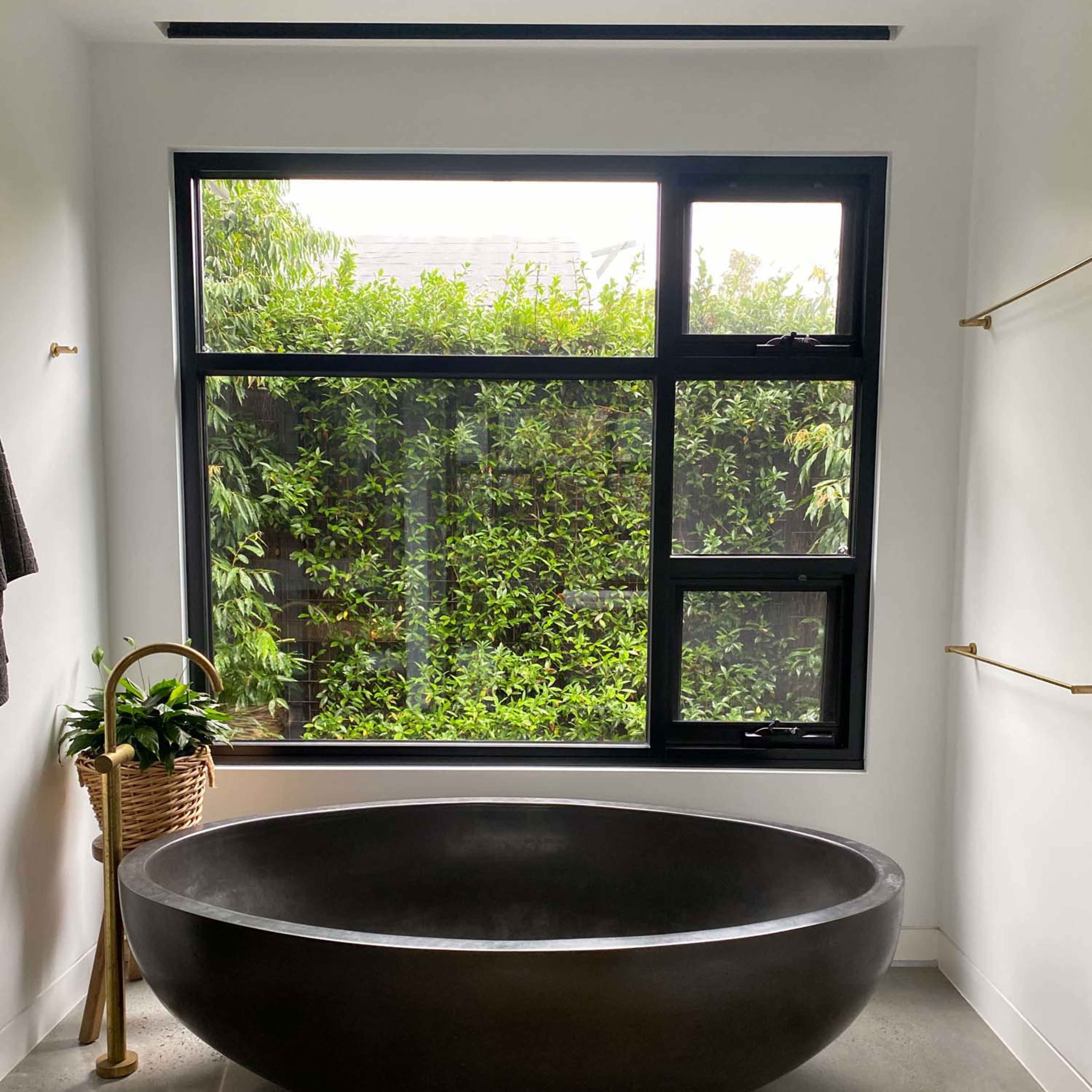 A modern bathroom featuring a black freestanding bathtub set against a large window with greenery outside.