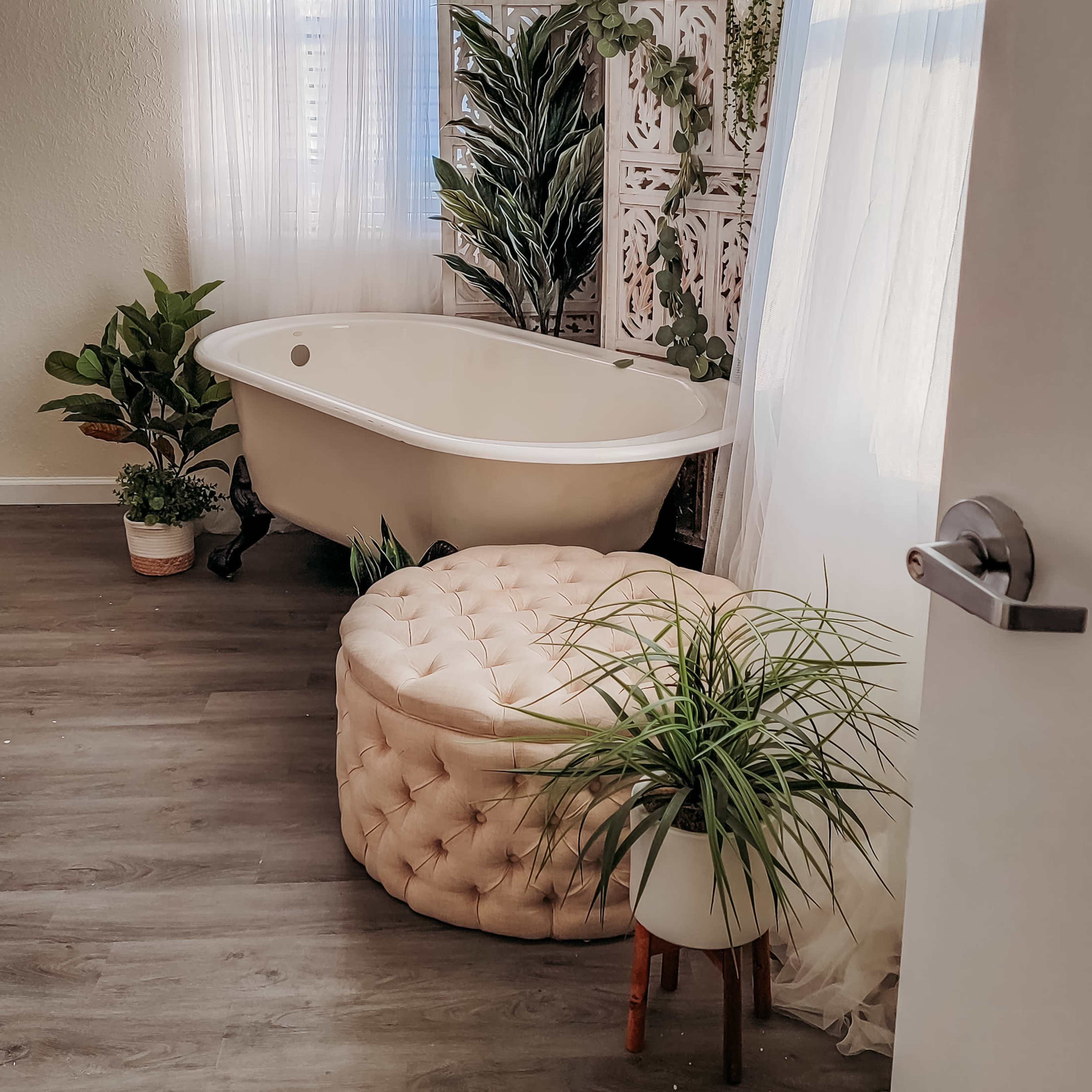 A bright bathroom featuring a freestanding bathtub, a large potted plant, a decorative wooden screen, and a tufted ottoman near a window.