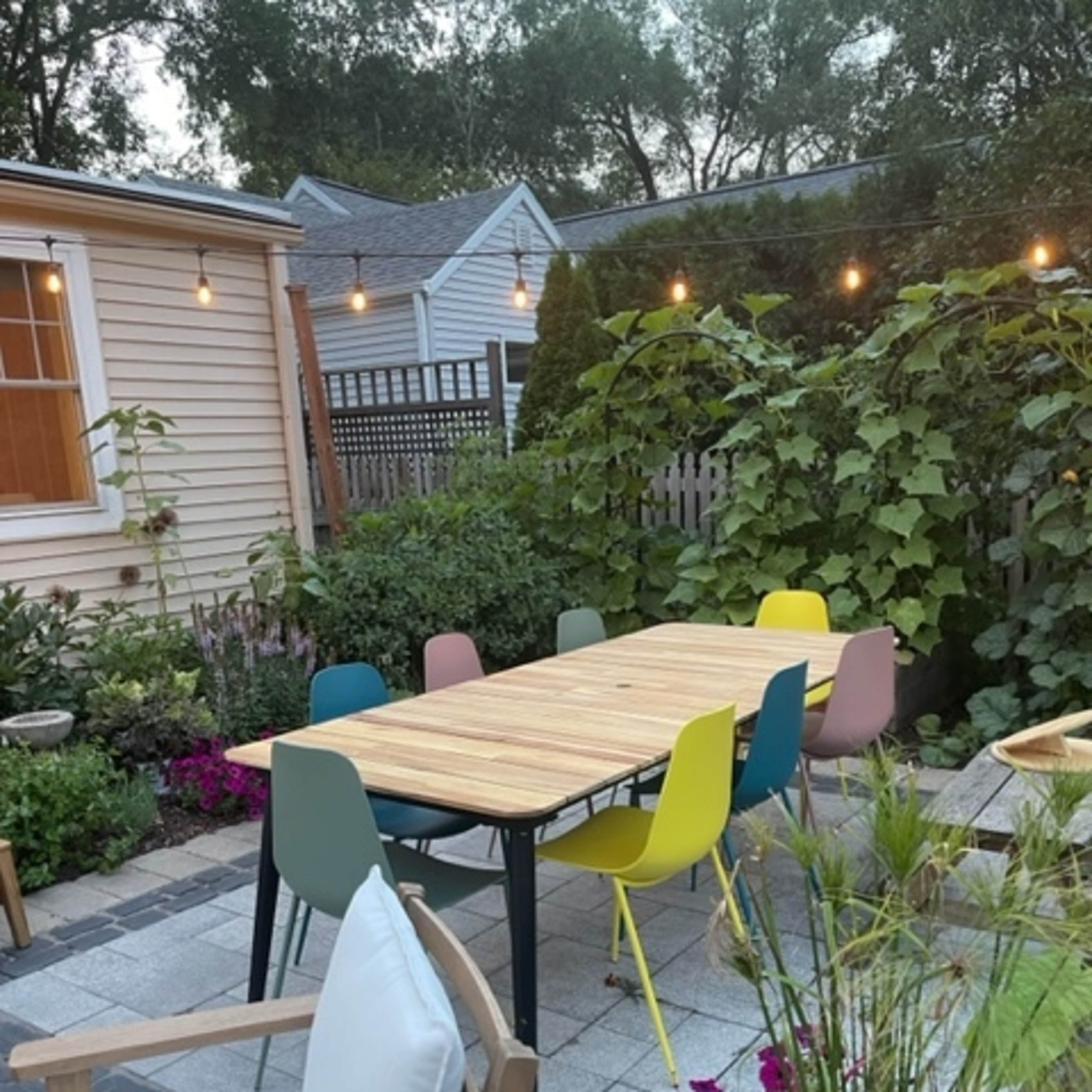 A wooden dining table surrounded by colorful chairs is set in a garden area with plants and flowers.