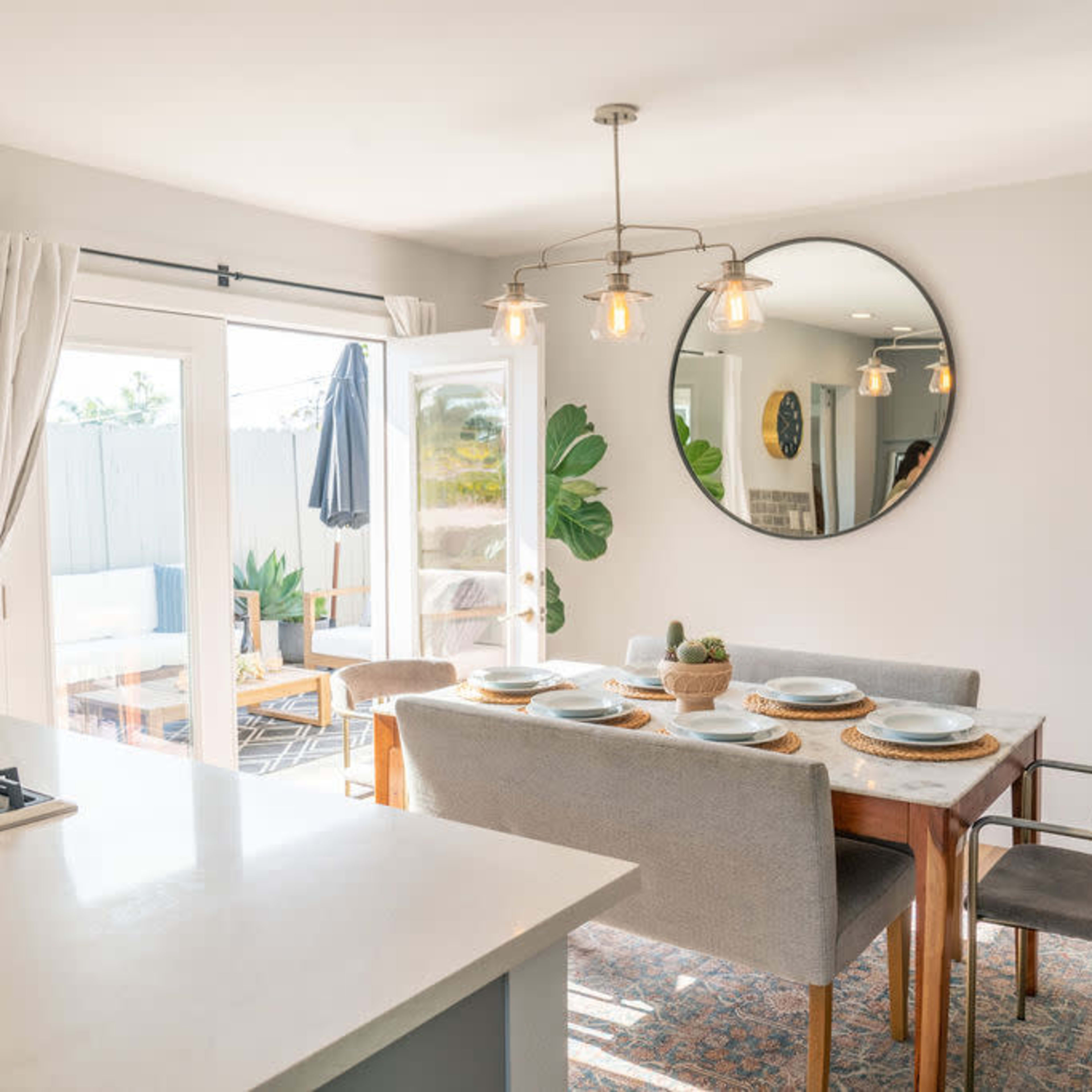 A dining area features a wooden table set with plates, surrounded by chairs, with large windows and a mirror reflecting the light.