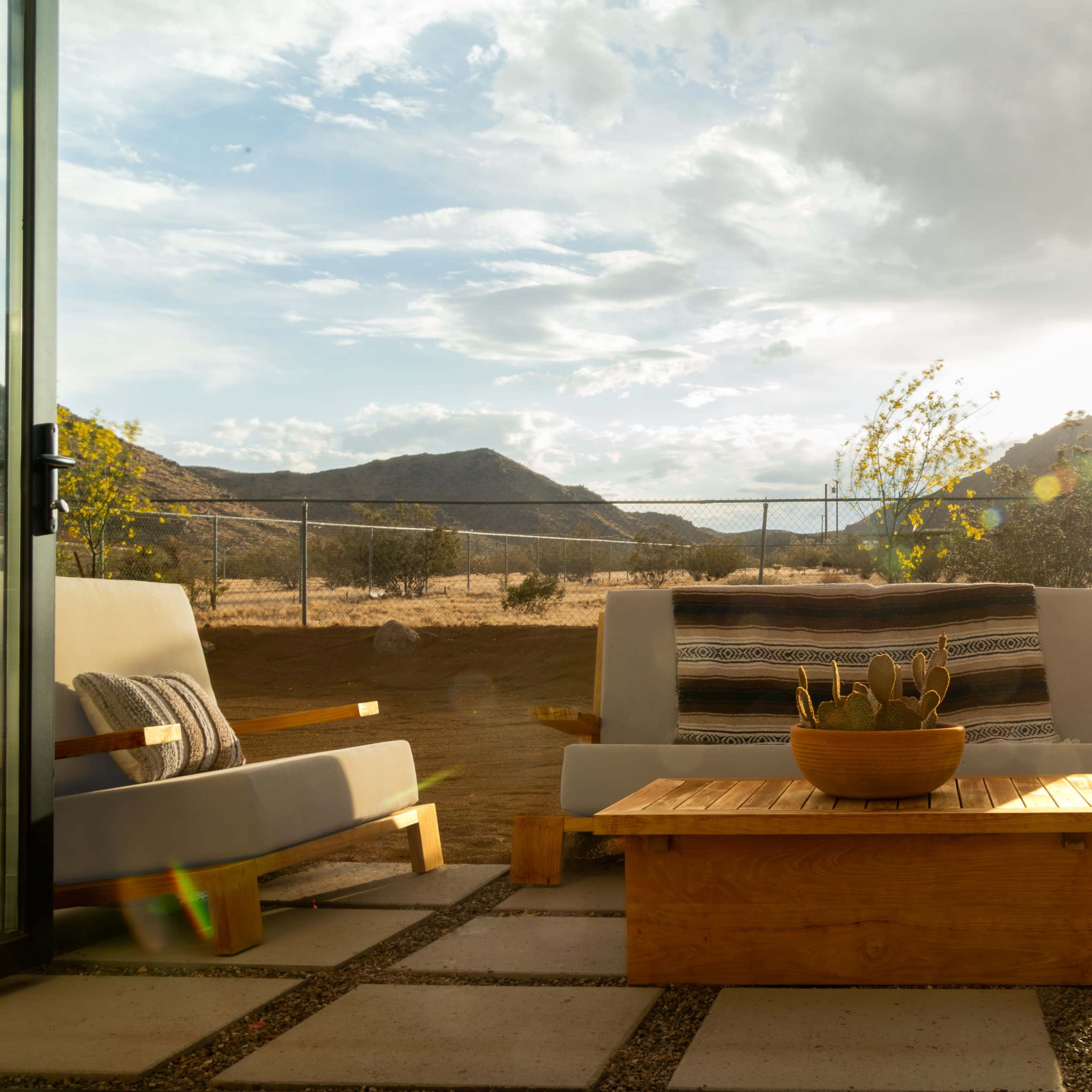 A cozy outdoor patio features two armchairs and a wooden coffee table, set against a backdrop of mountains and a rising sun.