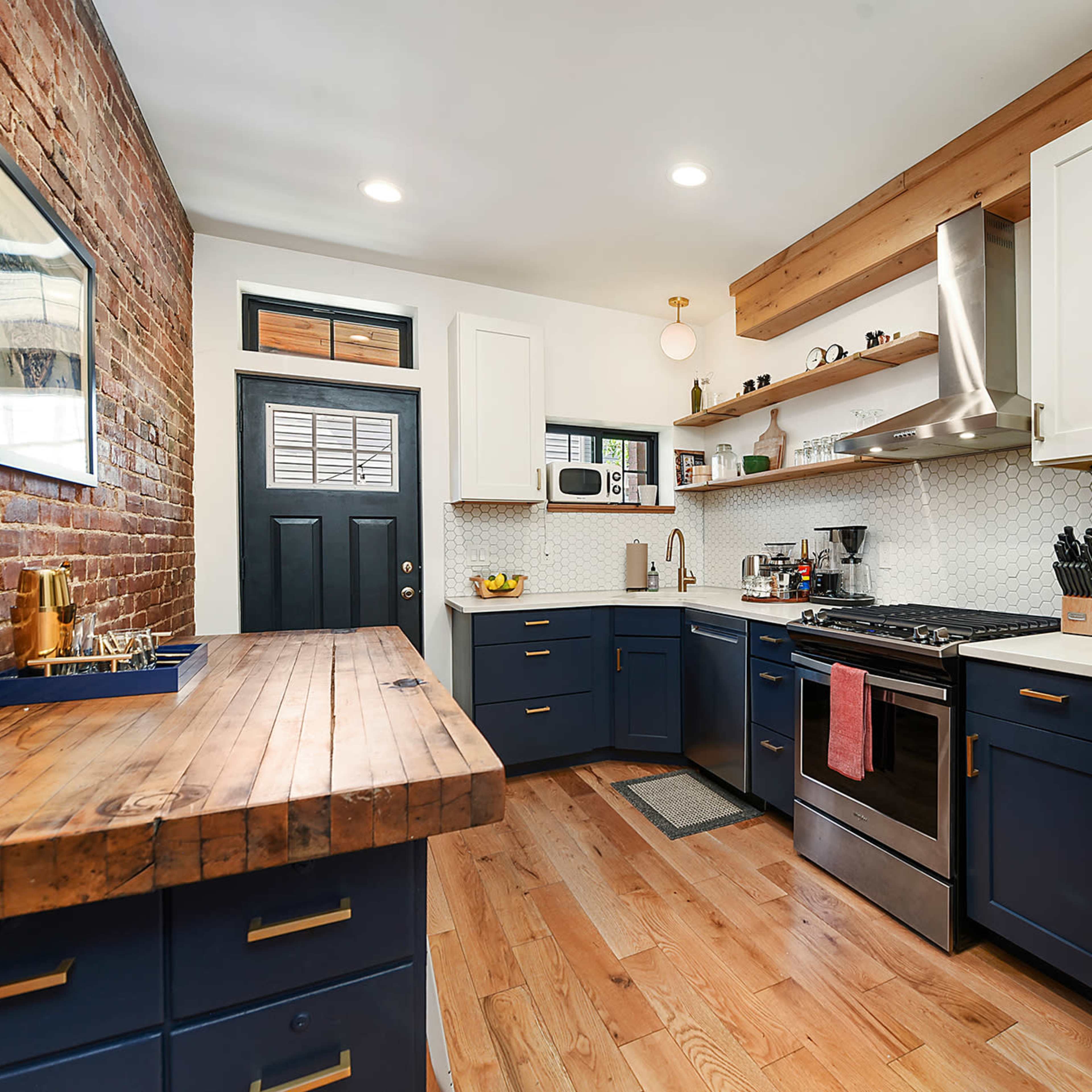 The kitchen features a combination of exposed brick and white cabinetry, with a wooden countertop, stainless steel appliances, and a door leading outside.