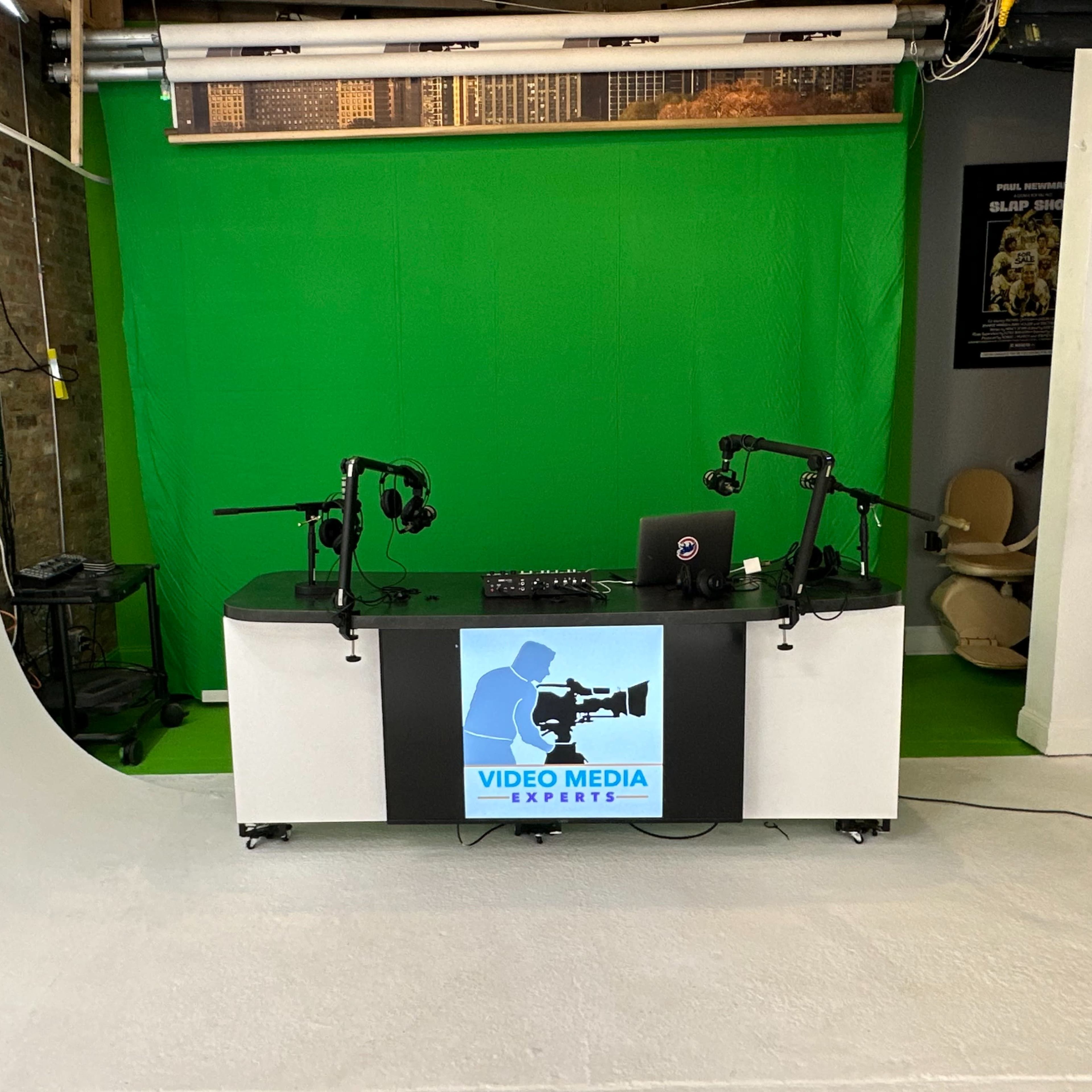 A video production studio features a green screen backdrop, a central black and white desk with a logo, and two microphone stands.