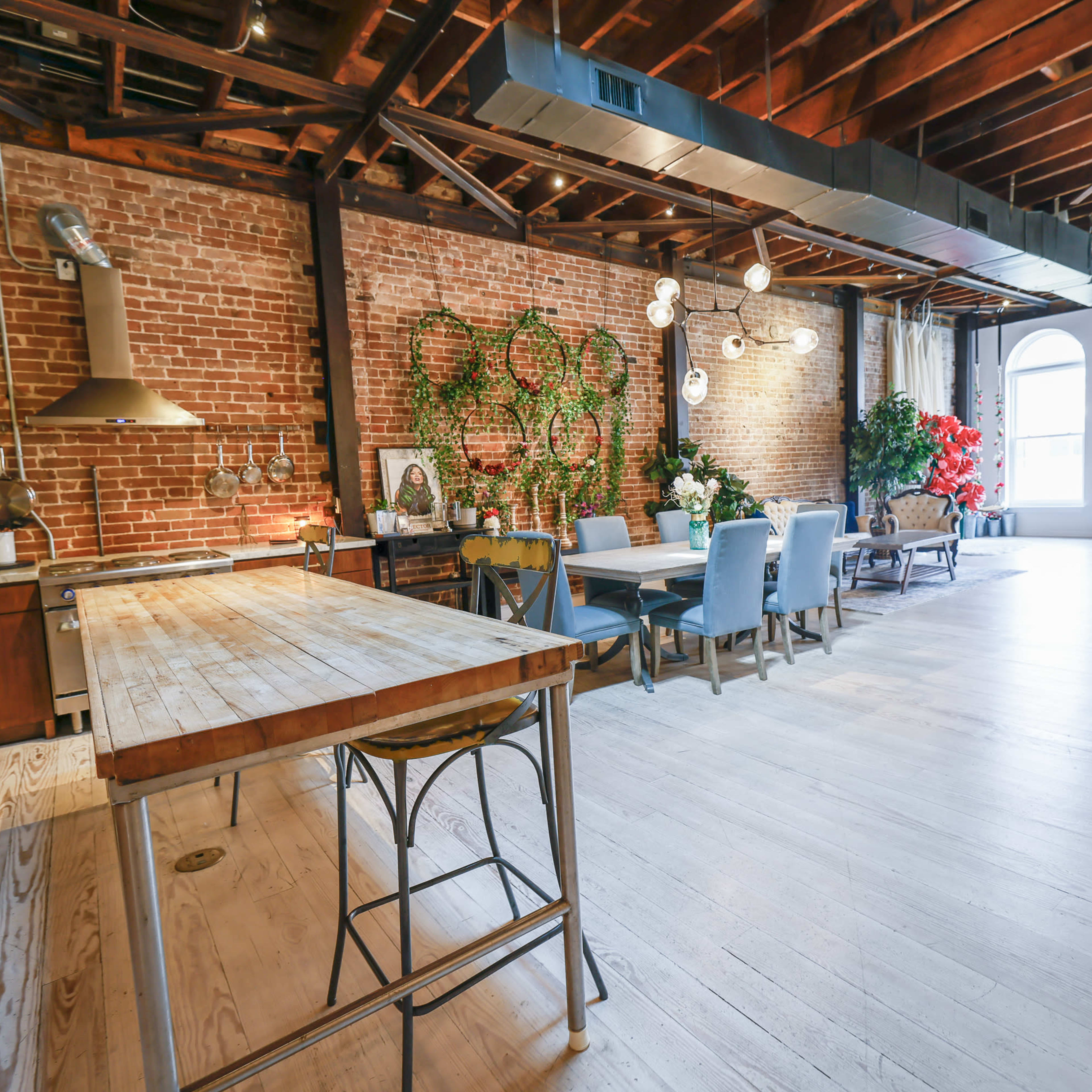 A spacious, modern loft-style kitchen and dining area featuring exposed brick walls, wooden beams, and a large wooden table surrounded by chairs.