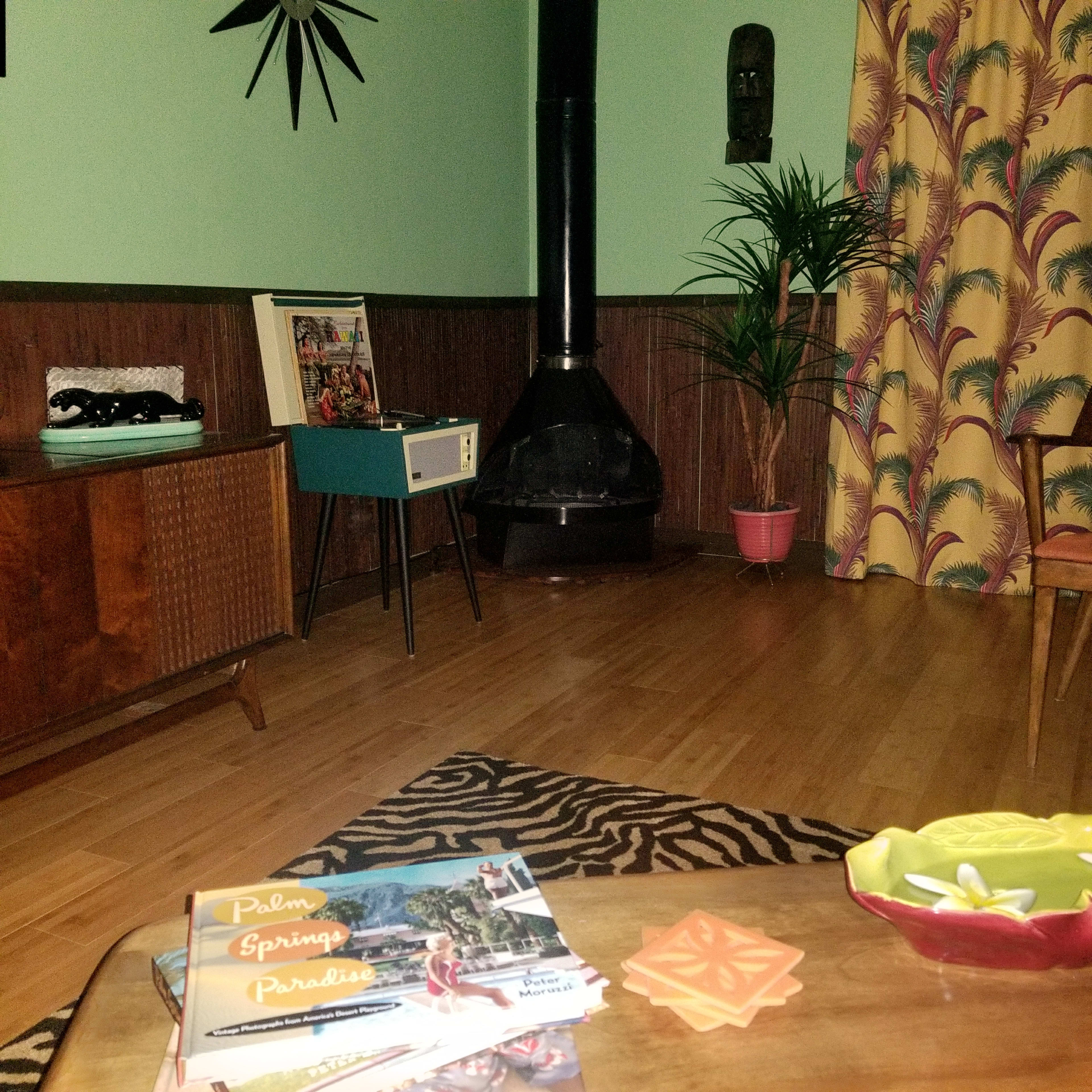 A mid-century modern living room featuring a vintage record player, a plant, and patterned curtains, with magazines displayed on a coffee table.