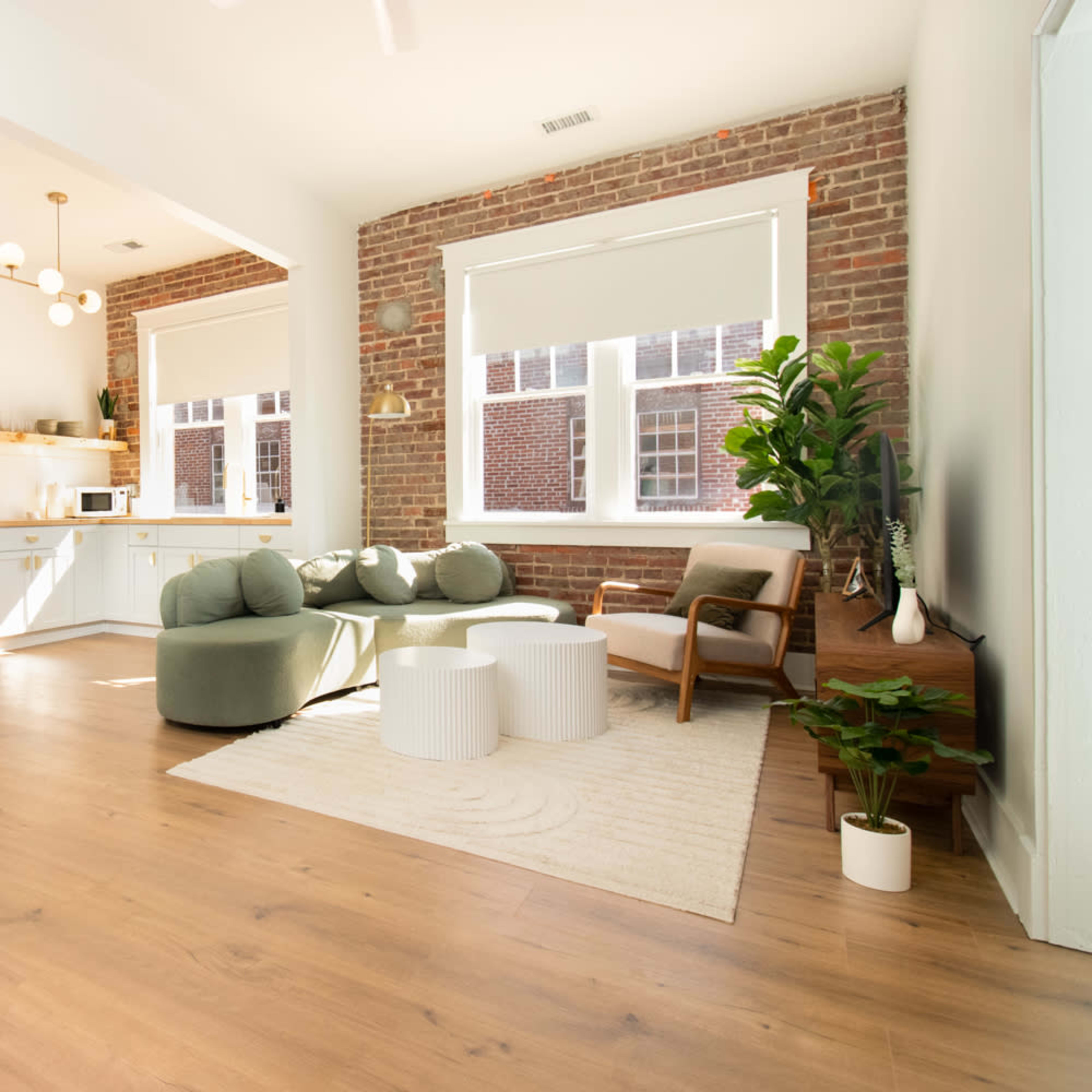 A modern living space with a kitchen area, featuring a seating arrangement of round chairs and a coffee table, alongside large windows and exposed brick walls.