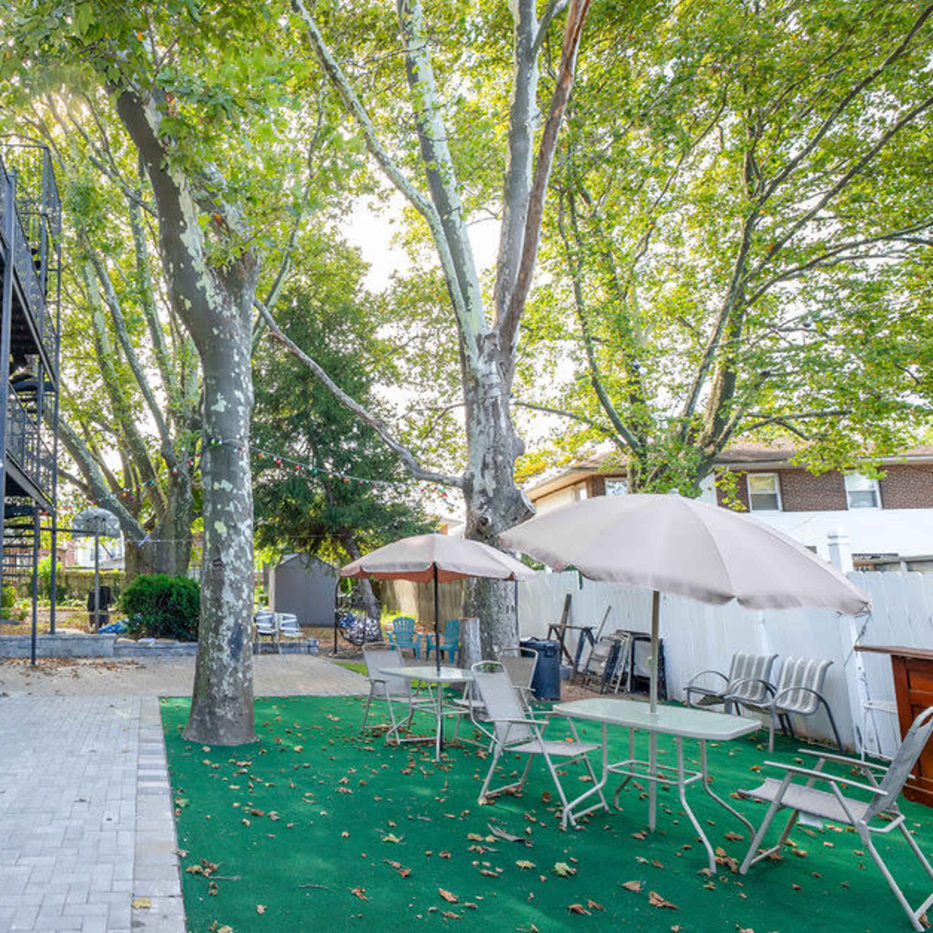 A backyard area with green turf, patio furniture under umbrellas, and a two-story building in the background.