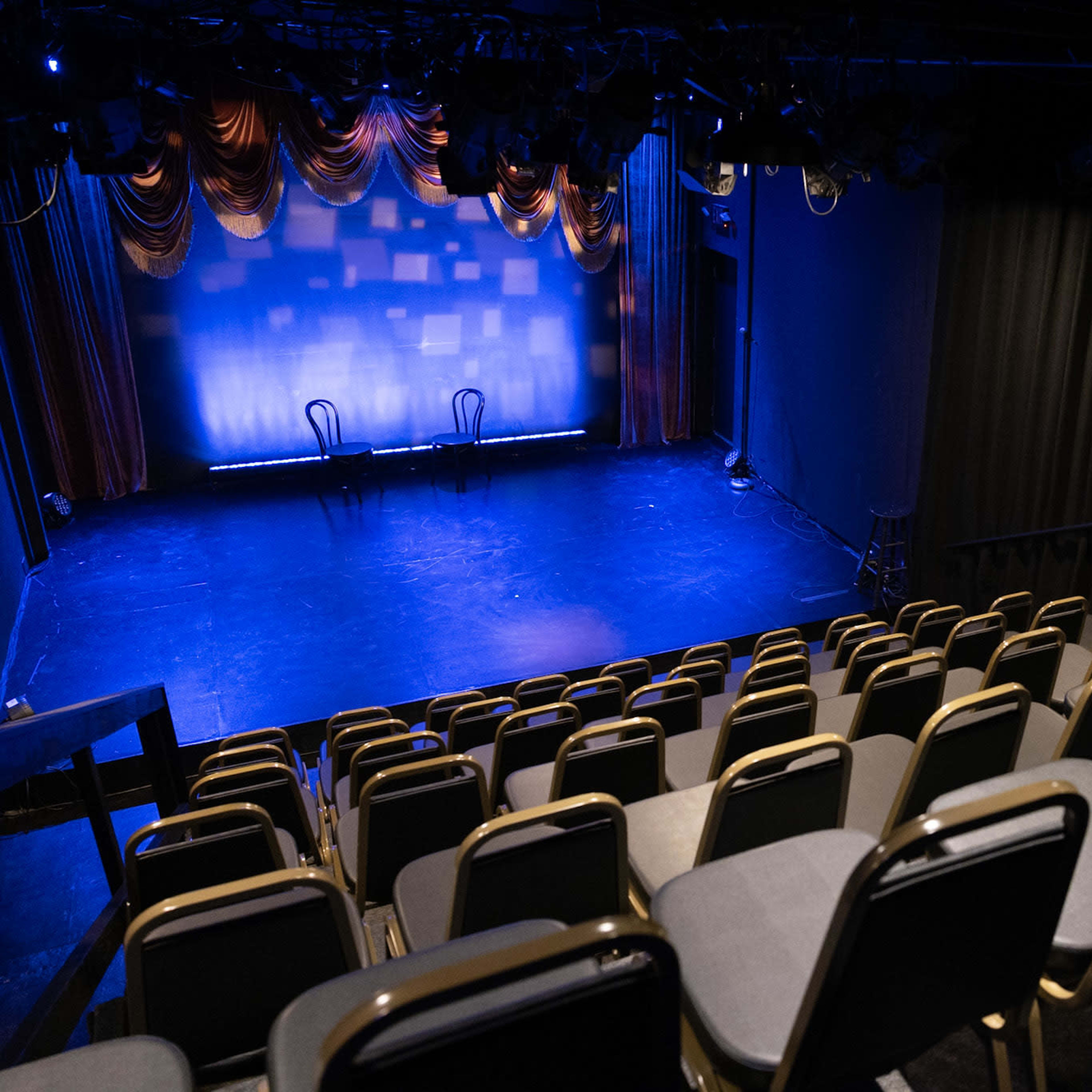 A small theater stage with two chairs on it, surrounded by empty rows of seating and dramatic curtains.