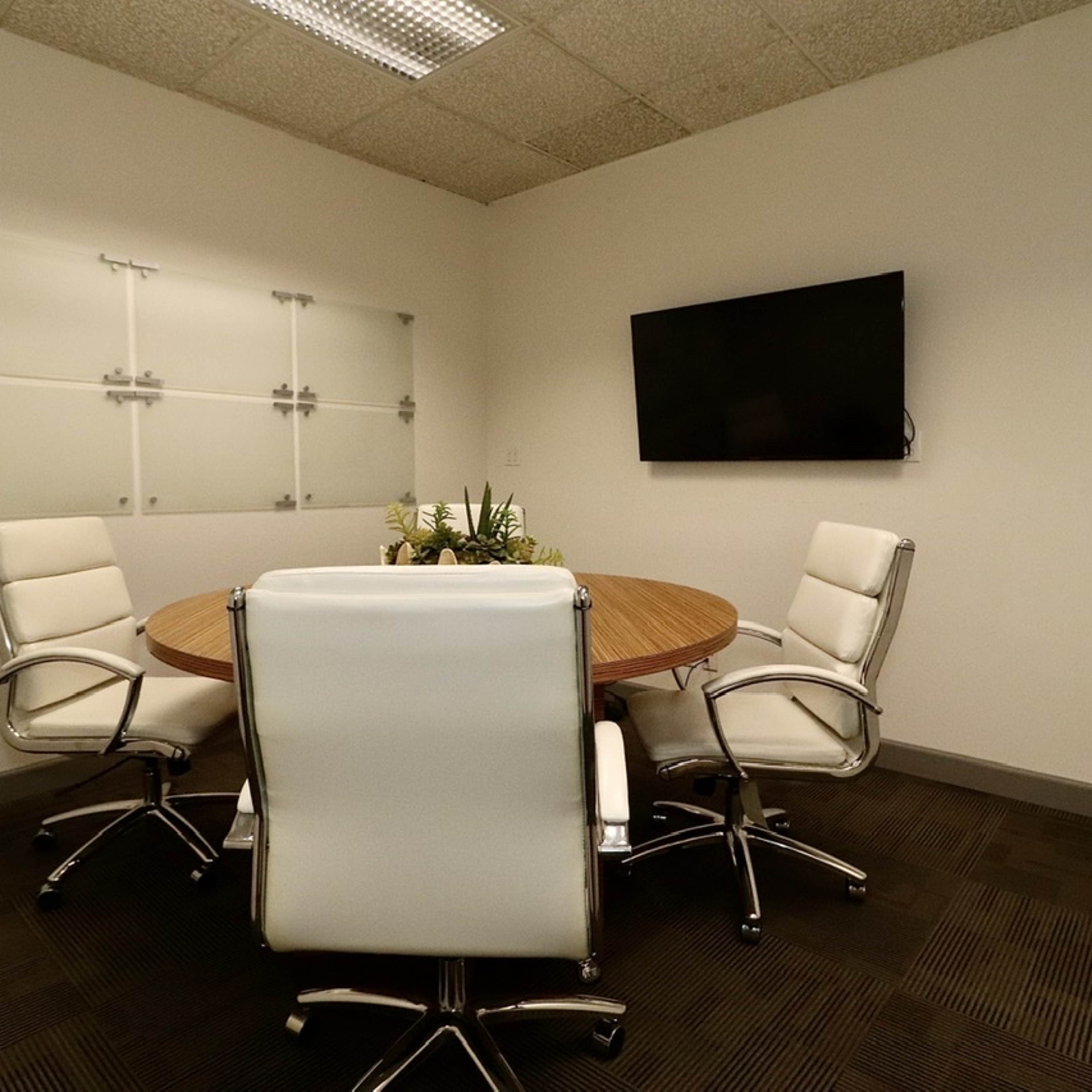 A small conference room features a round wooden table surrounded by four white leather chairs, with a mounted television and a glass whiteboard on the wall.