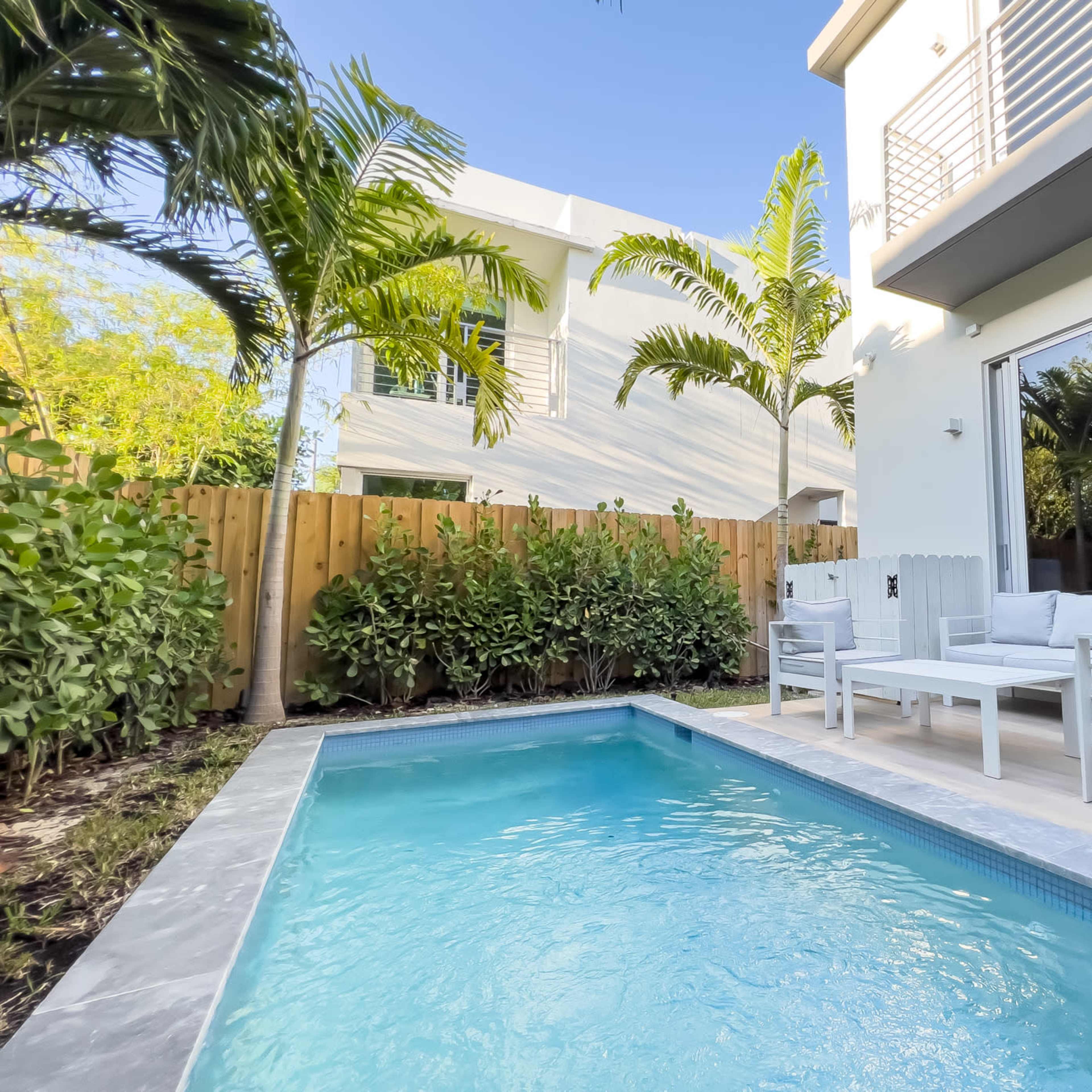 A small private pool surrounded by a wooden deck and tropical plants, adjacent to a modern house with large windows.