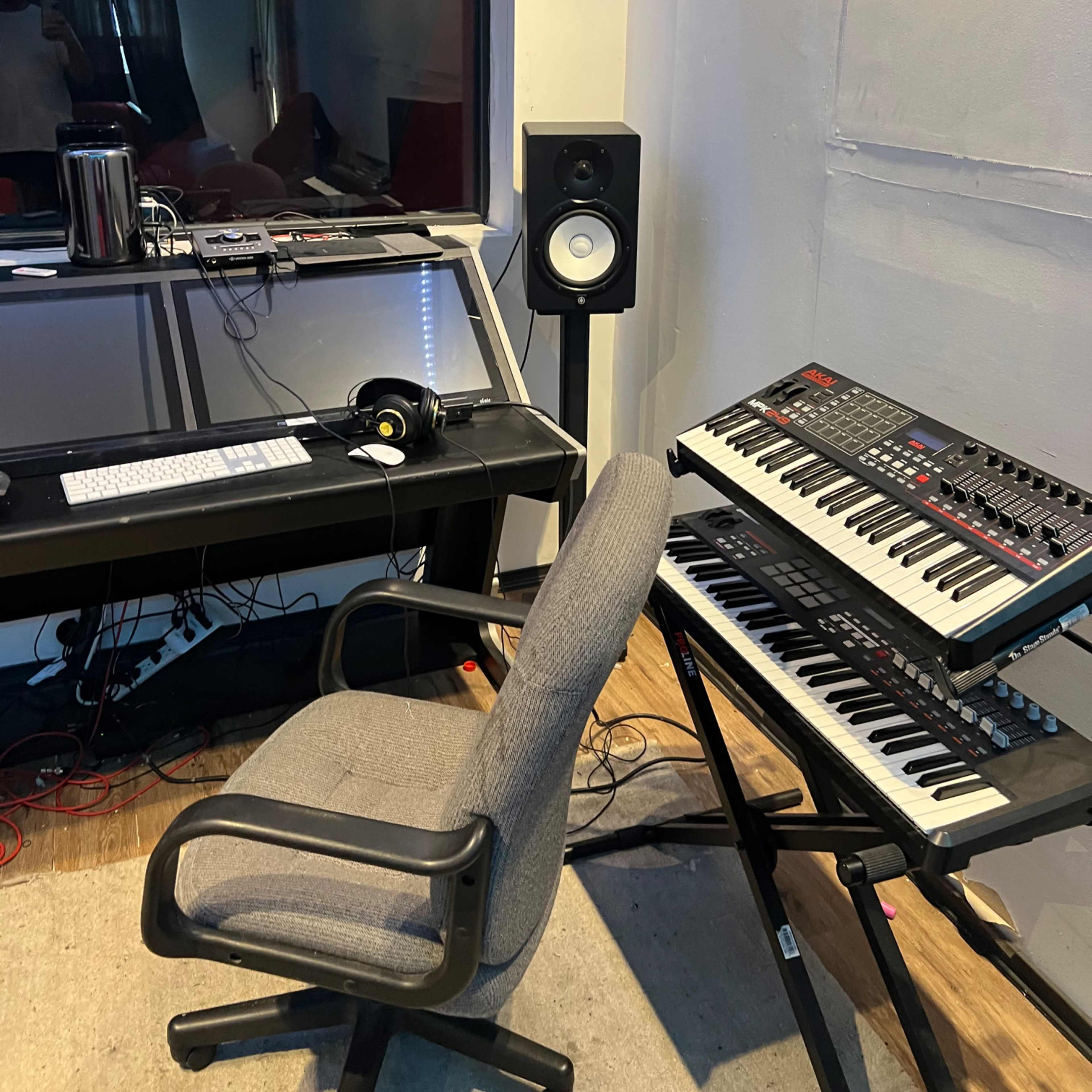 A music production studio features a computer desk, chair, audio monitors, and a synthesizer keyboard.