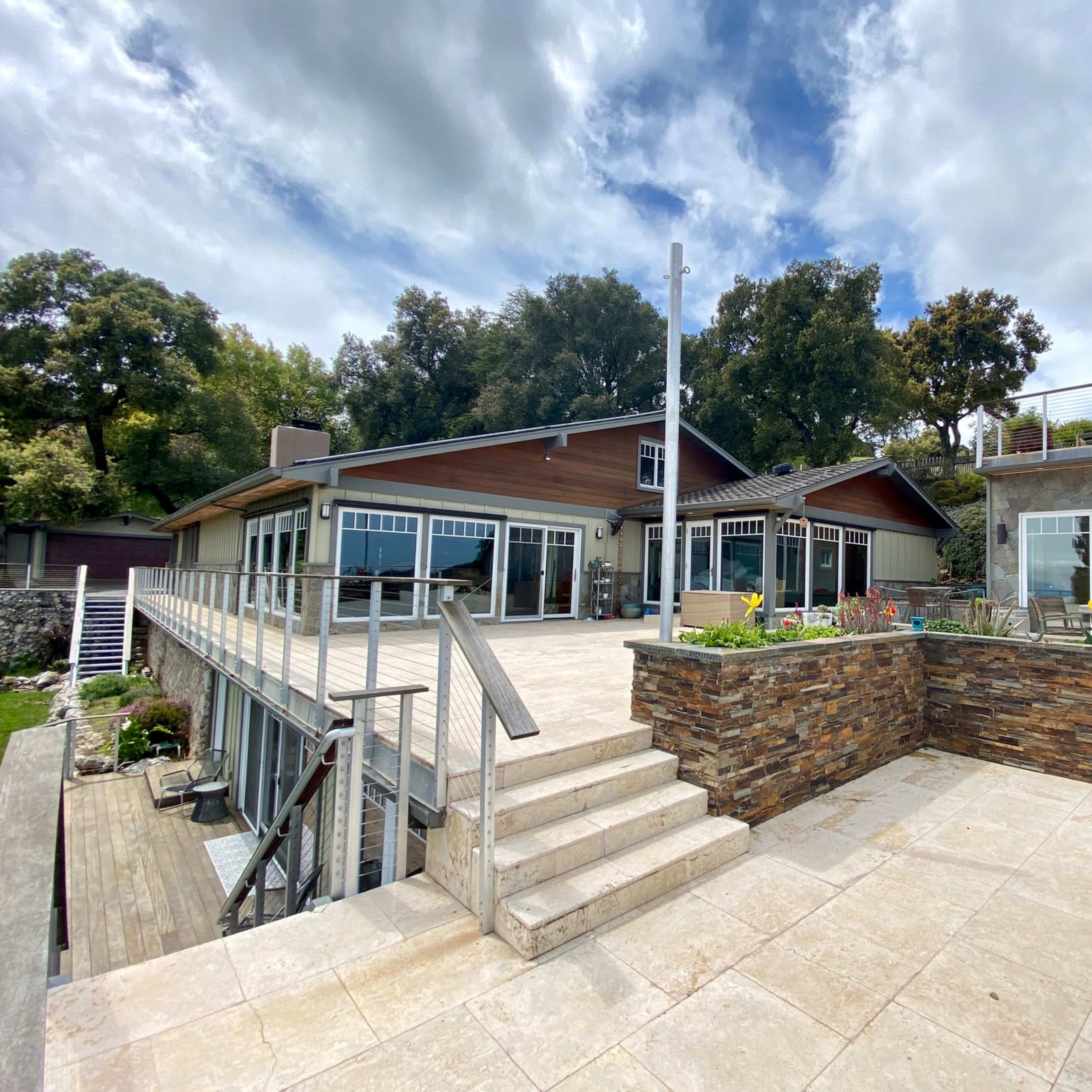 A modern two-story house with large windows, a stone patio, and steps leading down to a landscaped area surrounded by trees.