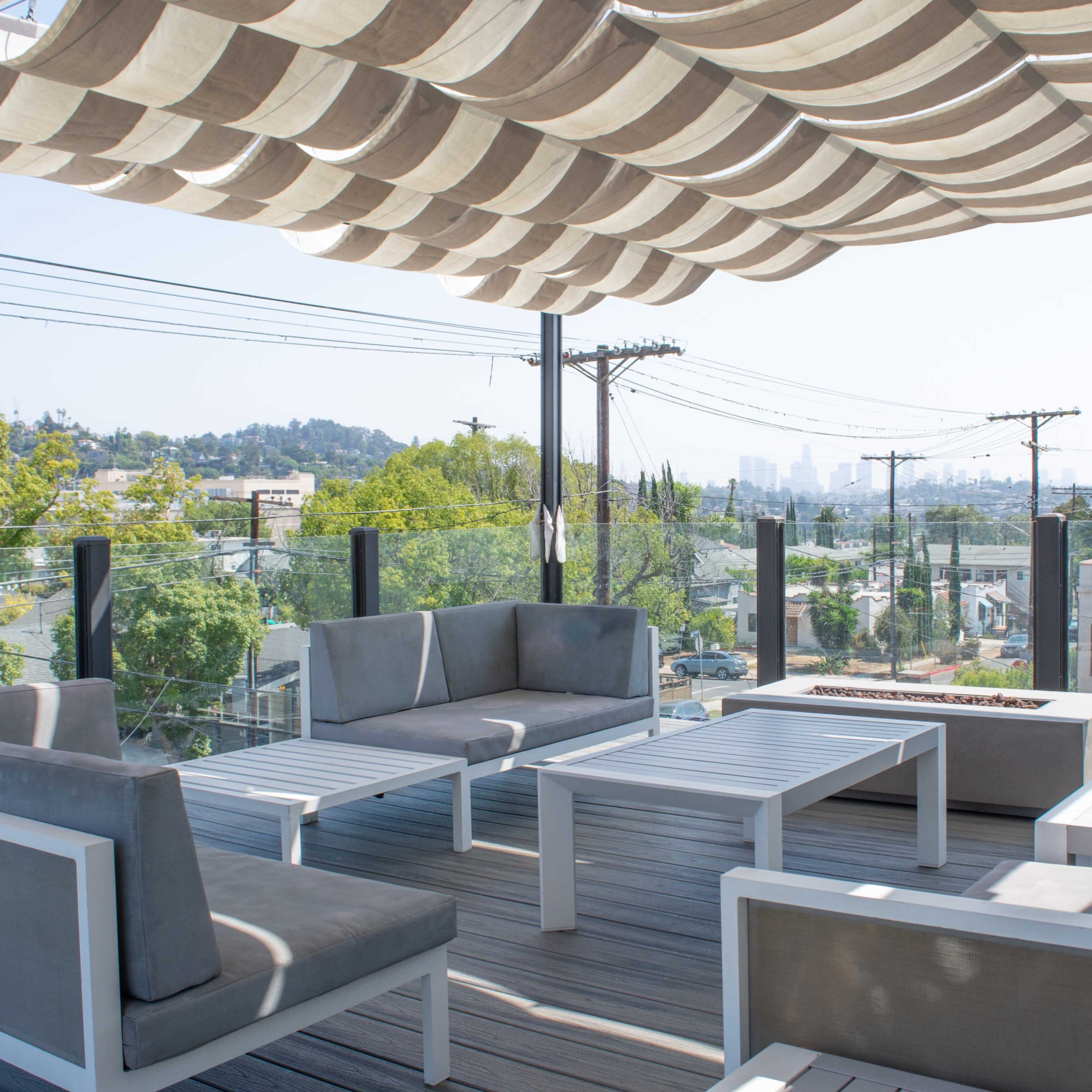 A modern rooftop terrace features gray seating arrangements and a white table under a striped awning, with a city skyline visible in the background.