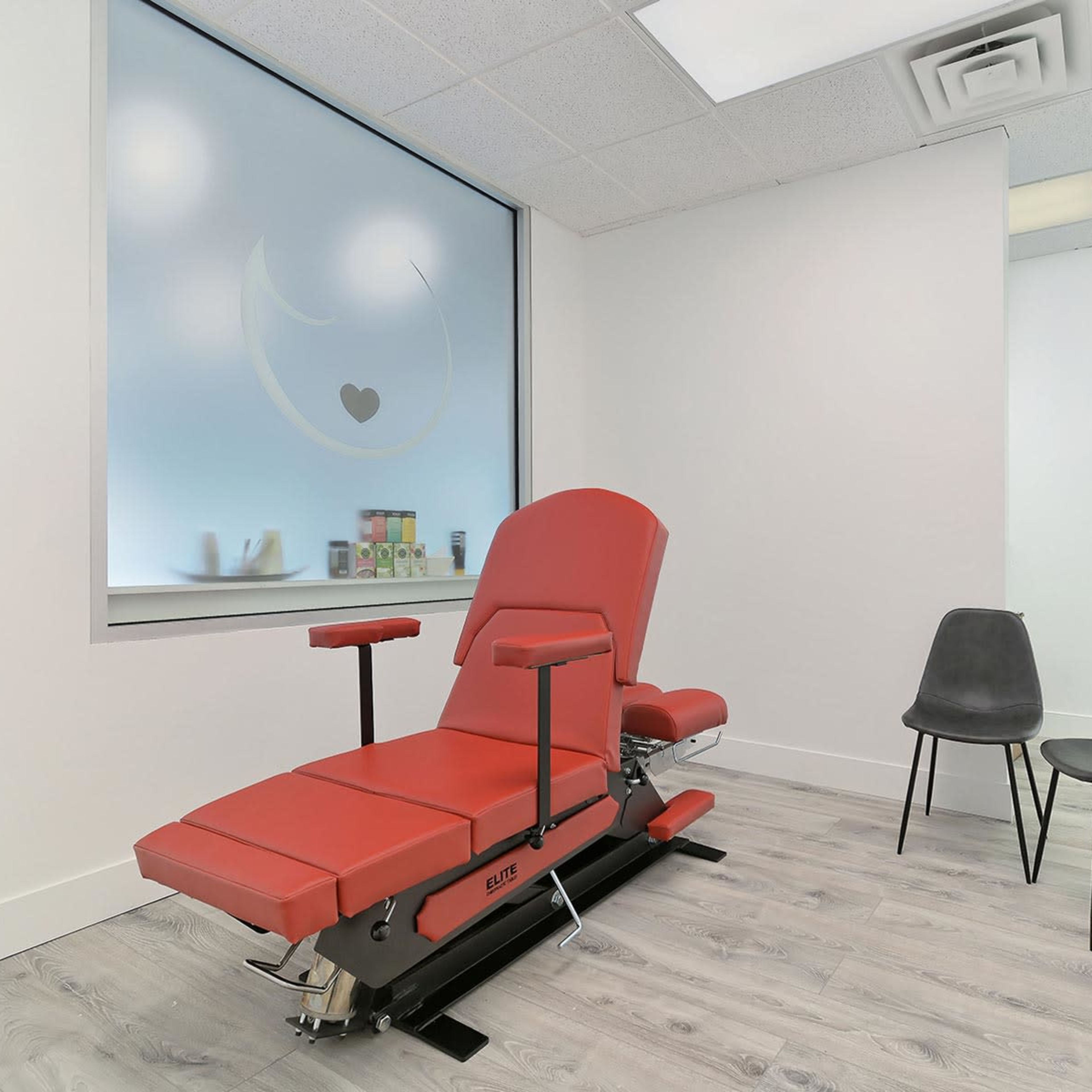 A minimalist treatment room equipped with a red examination chair and two black chairs against a light-colored wall.
