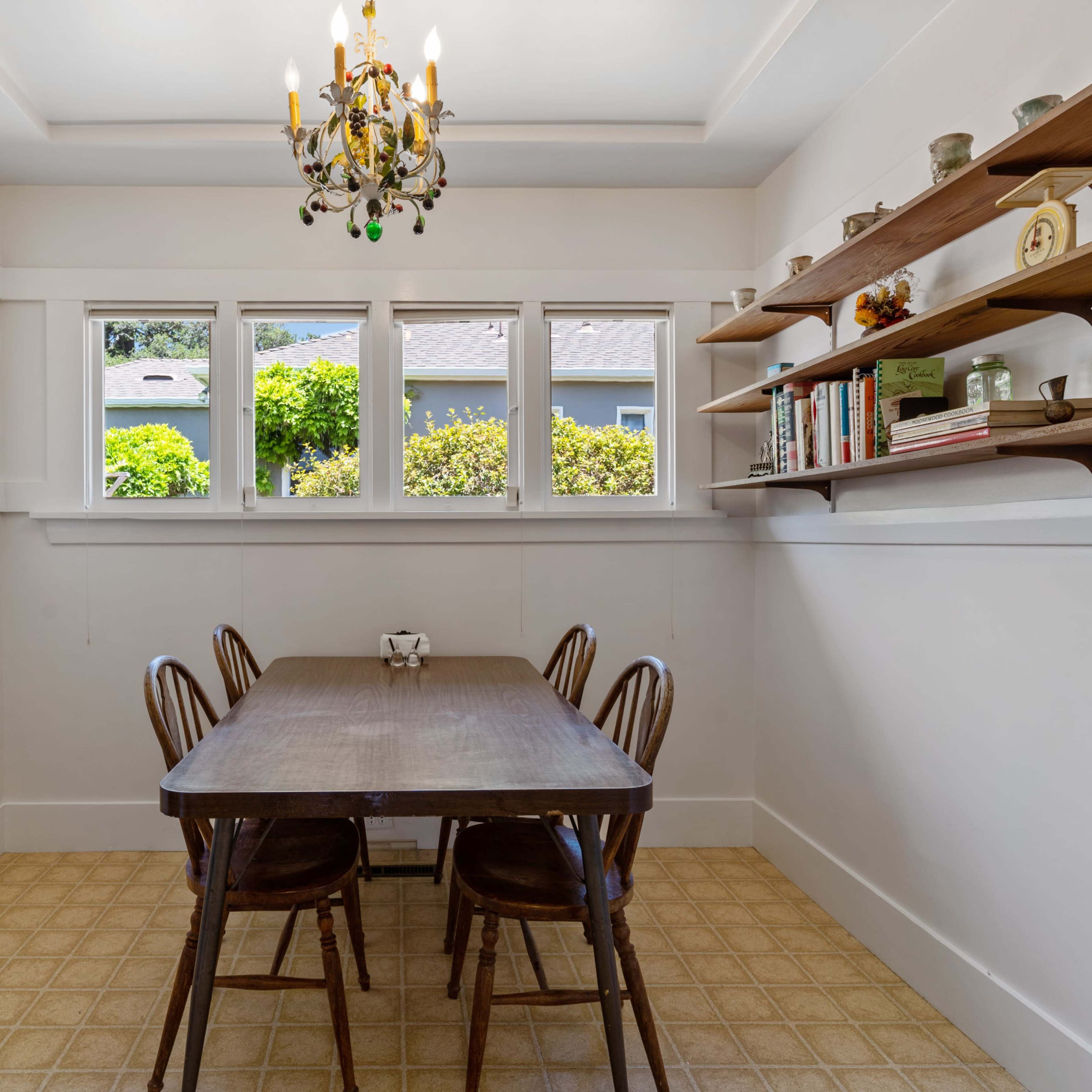 A dining area features a rectangular wooden table surrounded by four chairs, with a chandelier overhead and shelves displaying books and decorative items against the white walls.