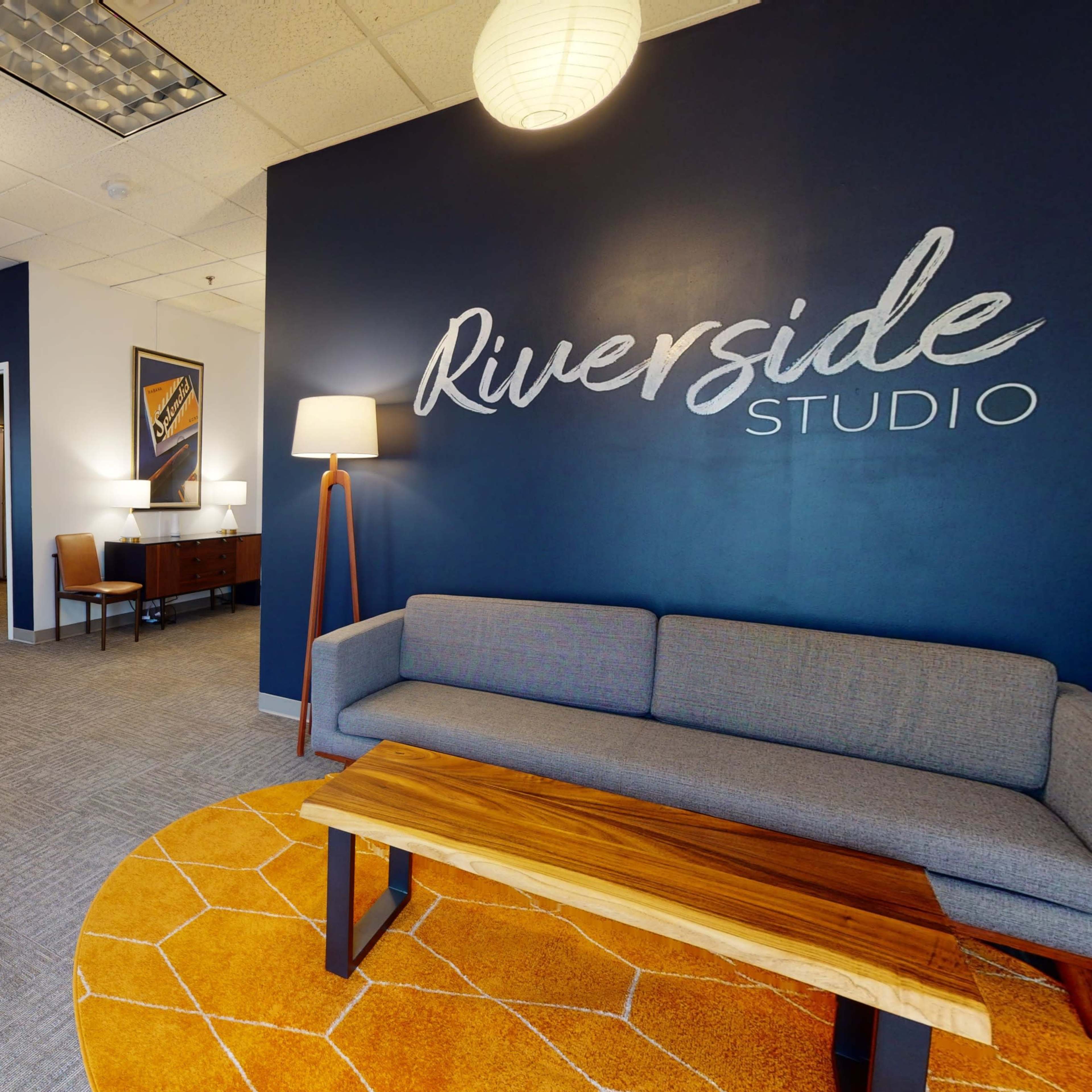 A modern studio interior featuring a gray couch, a wooden coffee table, a foosball table, and various seating arrangements against a dark blue accent wall with the words "Riverside Studio" painted on it.