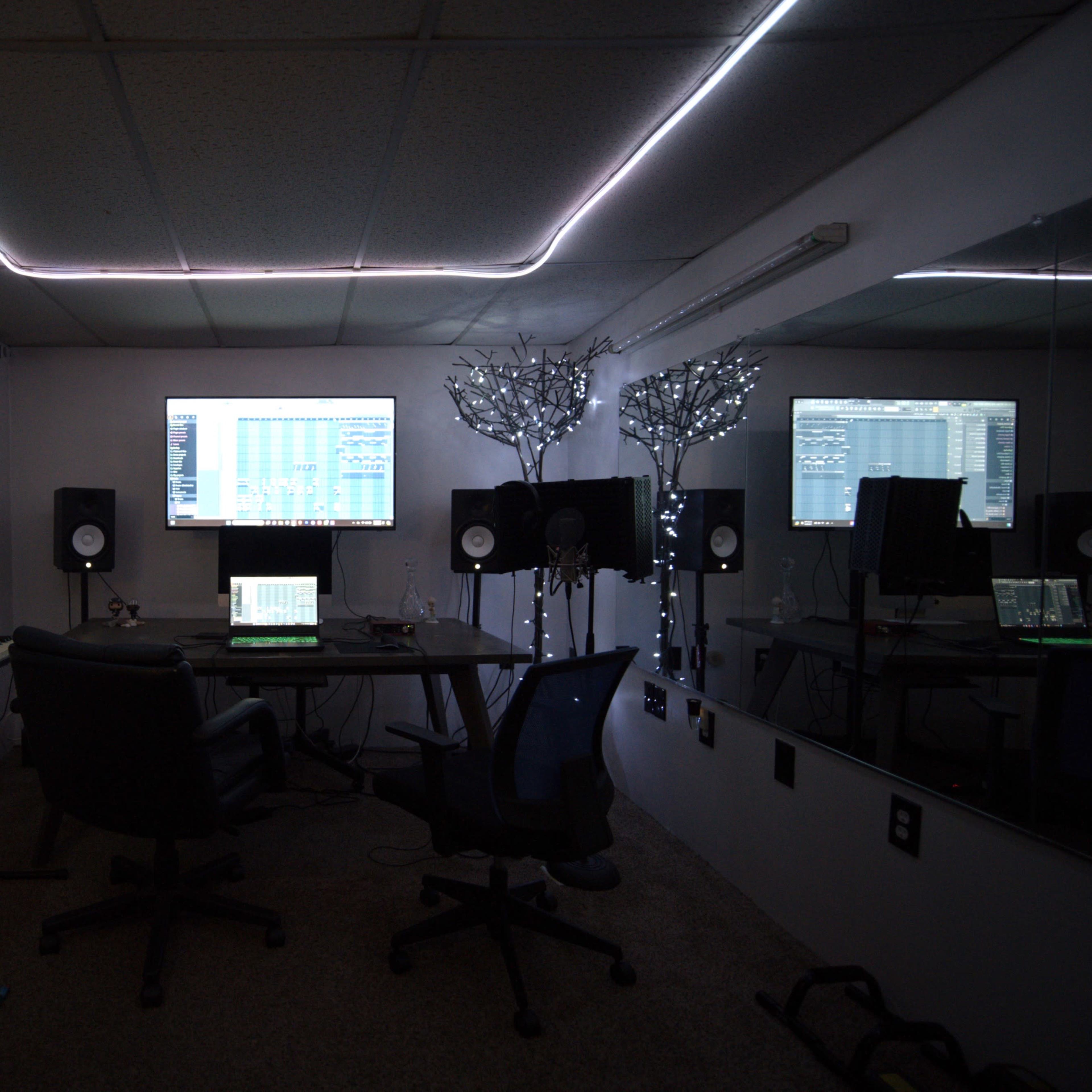 A music production studio featuring a desk with a computer, speakers, and a keyboard, alongside a large screen displaying audio software, all set against a backdrop of ambient lighting and mirrored walls.