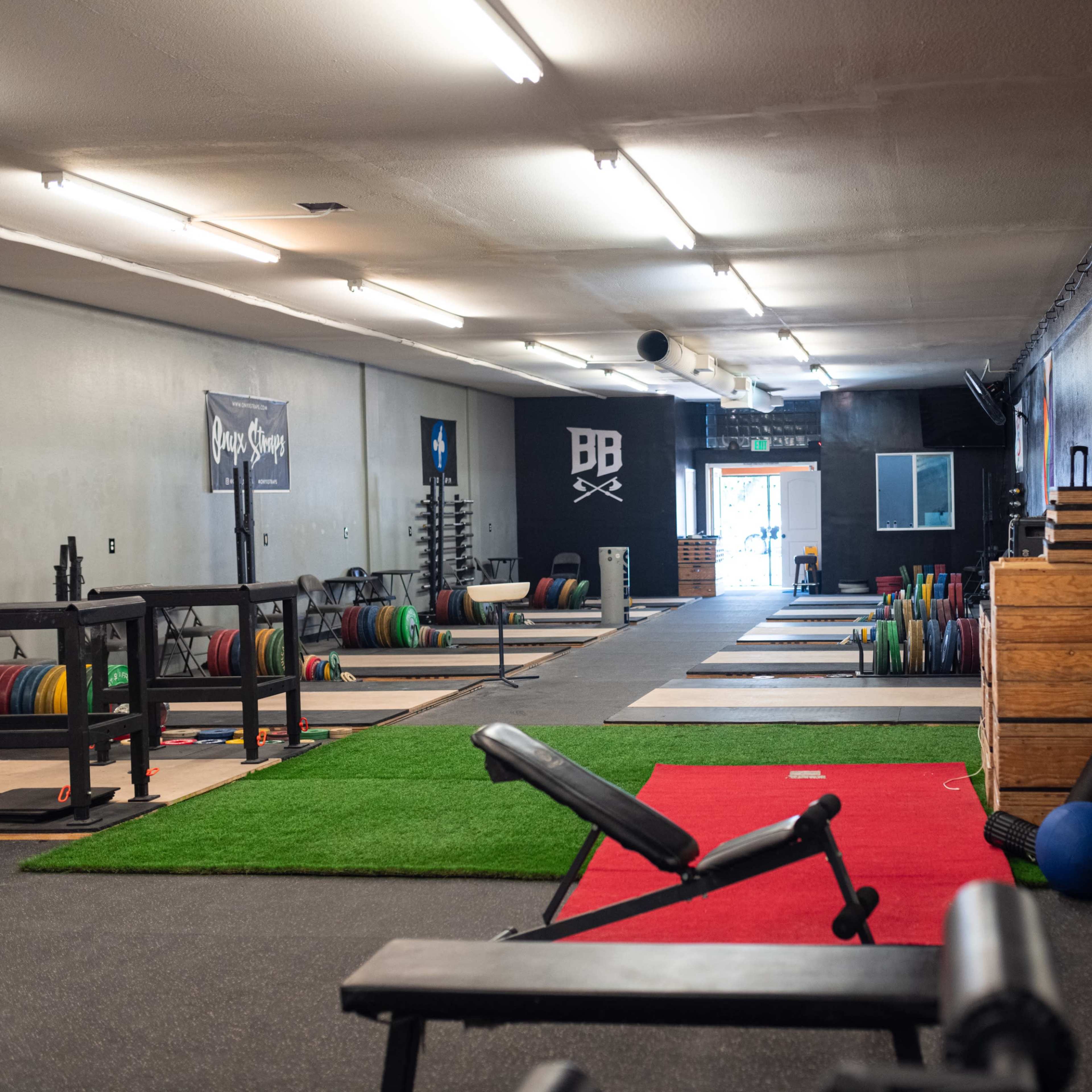 A spacious gym with weightlifting equipment, benches, and a variety of colorful plates arranged on racks along with a section of turf flooring.