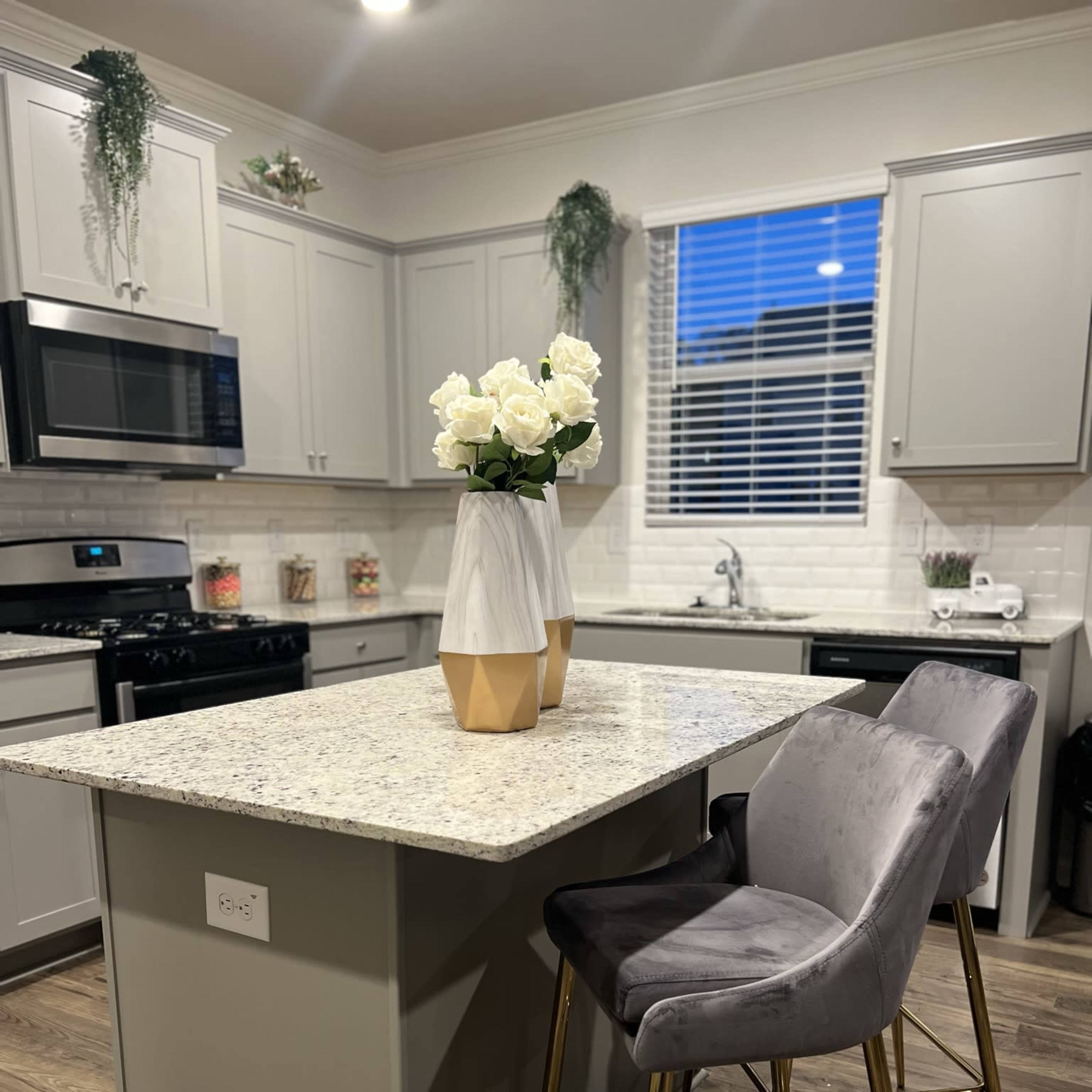 A modern kitchen features gray cabinetry, a granite island with a vase of white roses, and tall stools with gold legs.