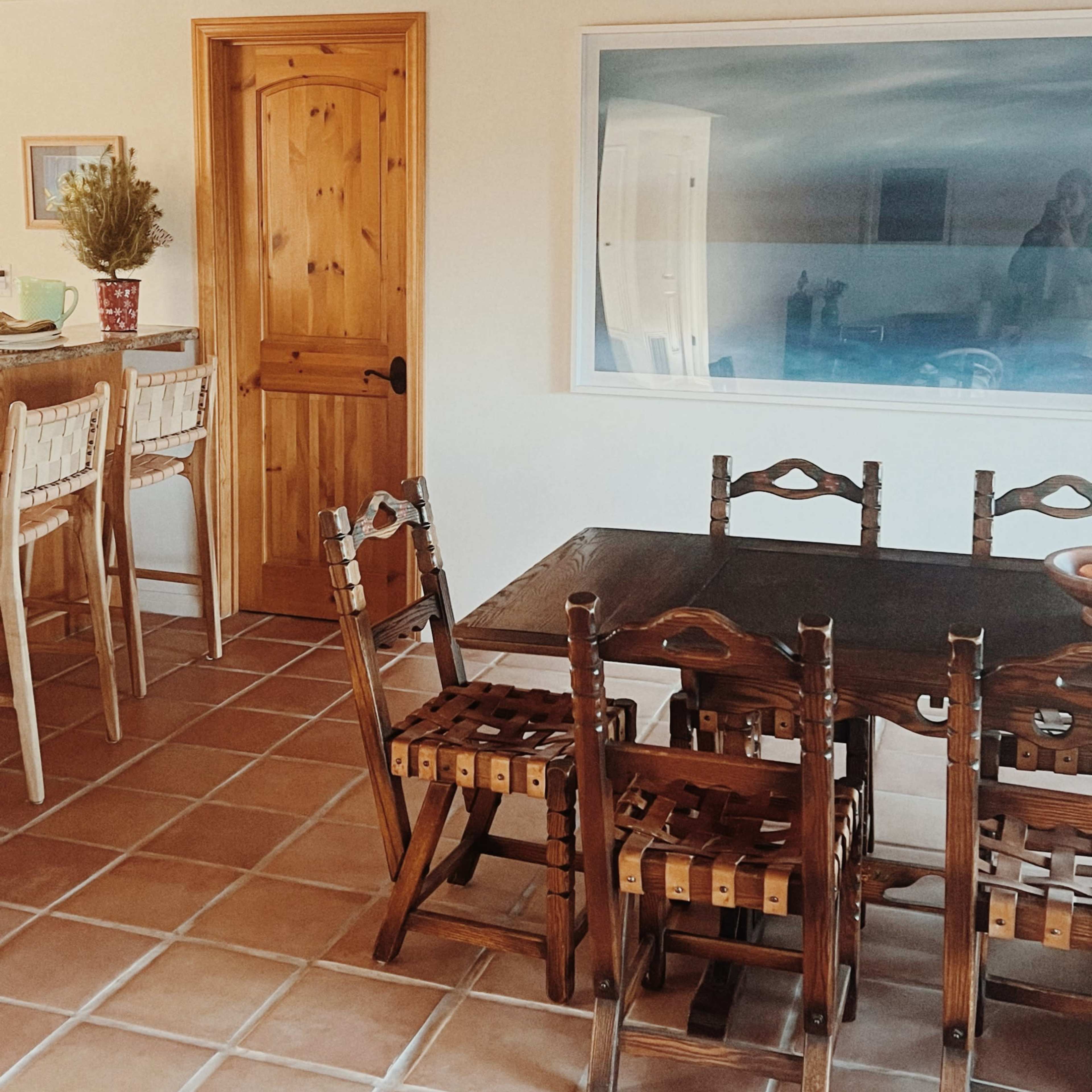 A dining area featuring a wooden table surrounded by four chairs, with bar stools positioned at a counter and a large painting on the wall.