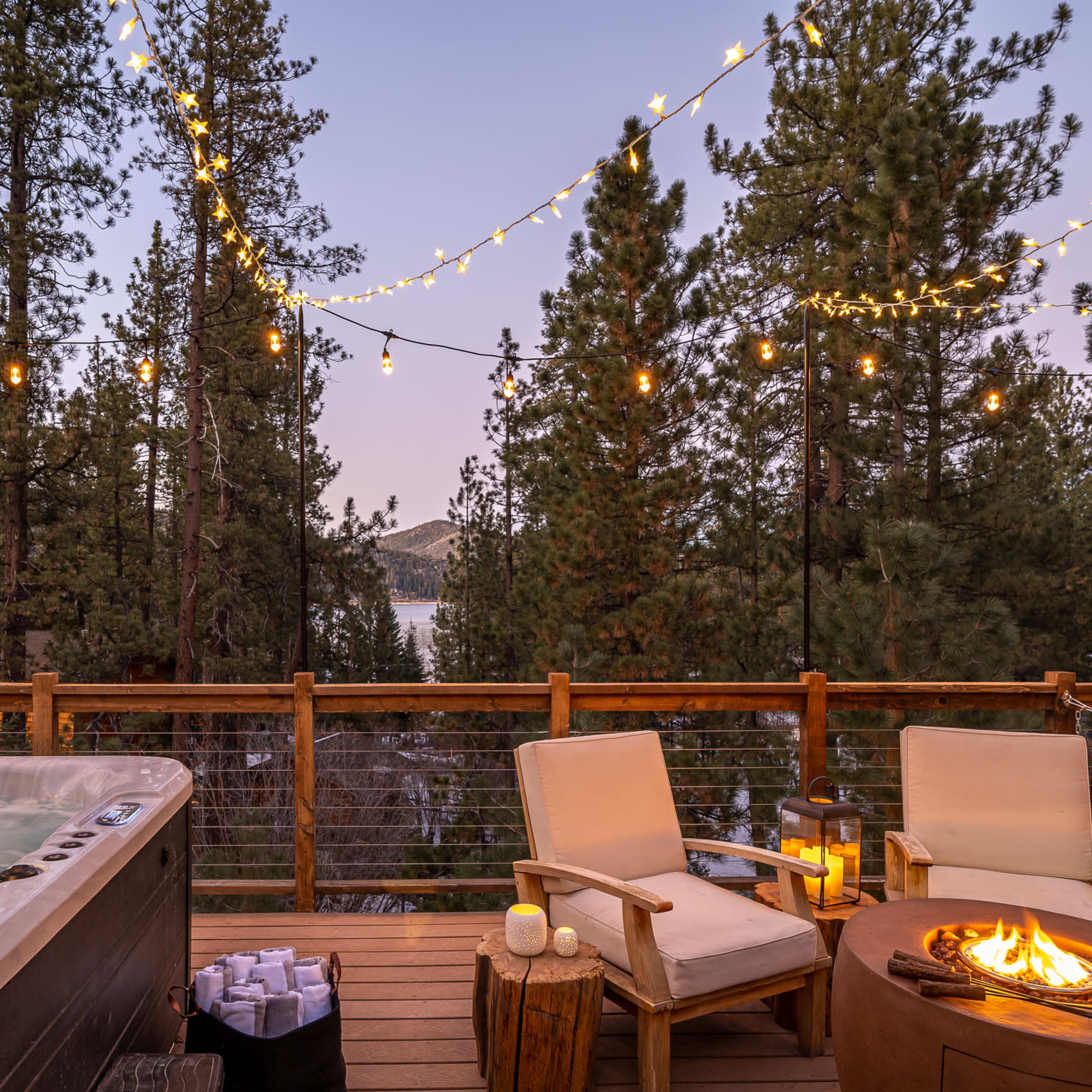 A wooden deck features a hot tub and two chairs near a fire pit, surrounded by tall trees and string lights, with a view of a lake in the distance.