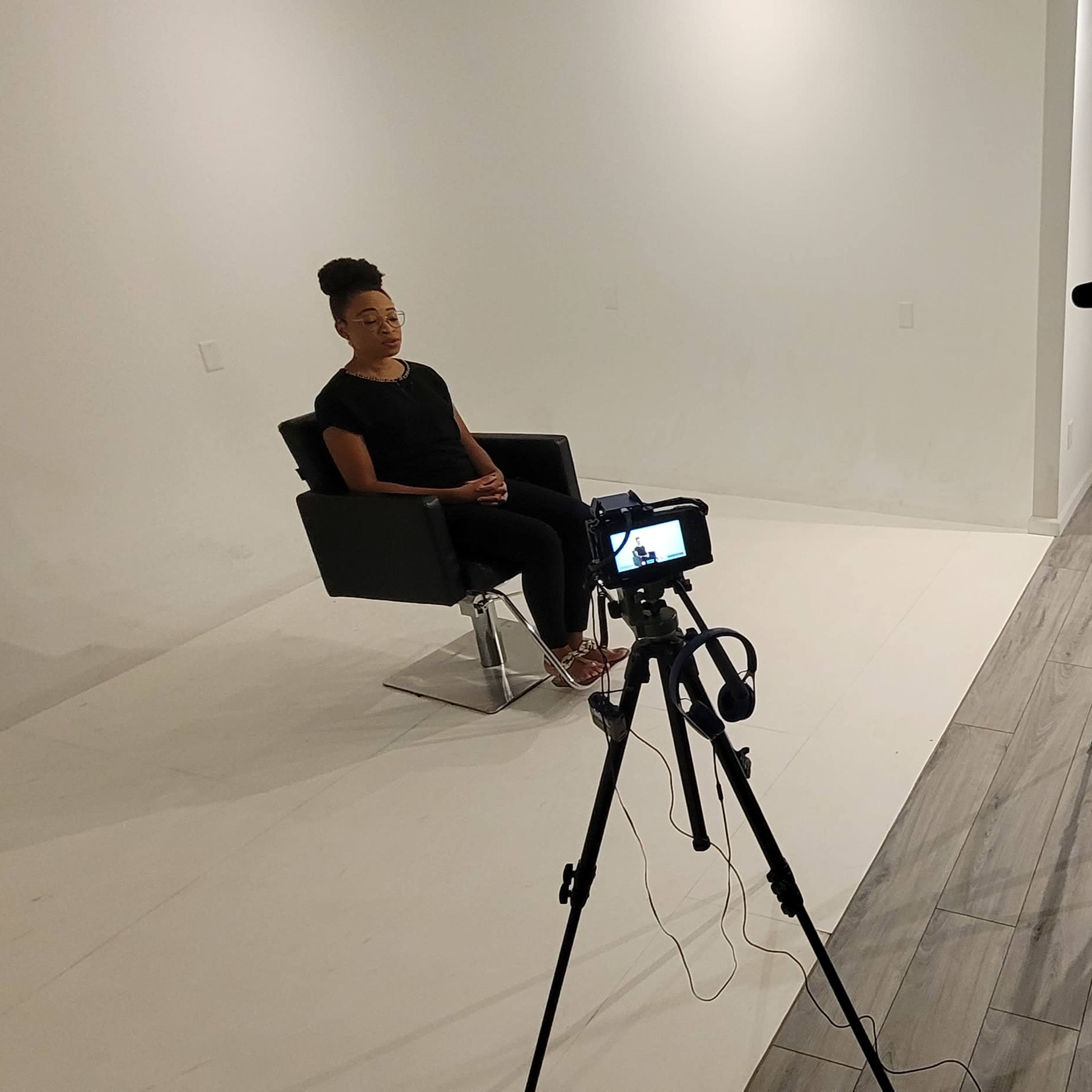 A person sits in a chair during an interview while another person operates a camera, and a third person stands in the background holding a clipboard.