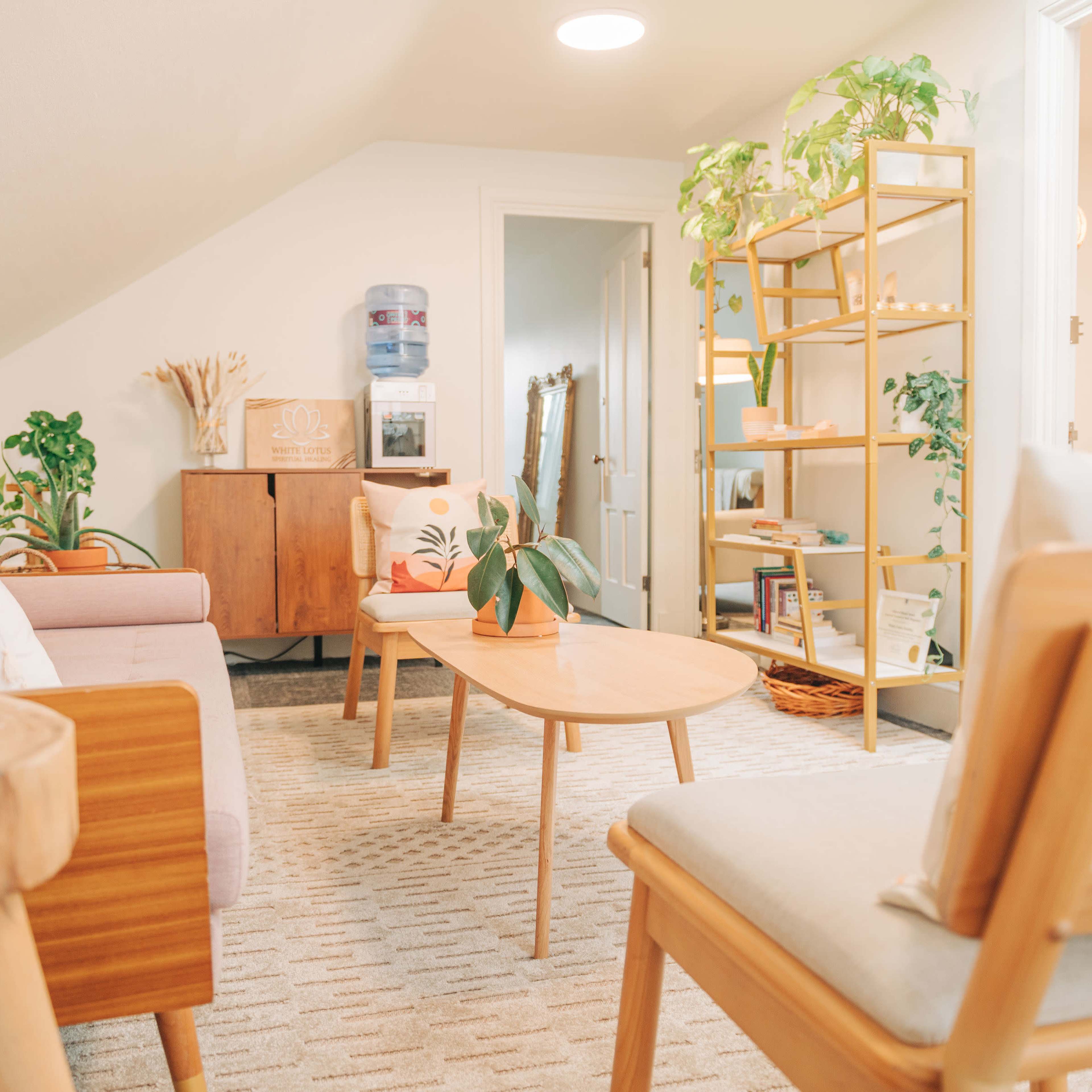 A cozy attic living space featuring two chairs, a wooden table, a sofa, and shelves adorned with plants and decorative items.