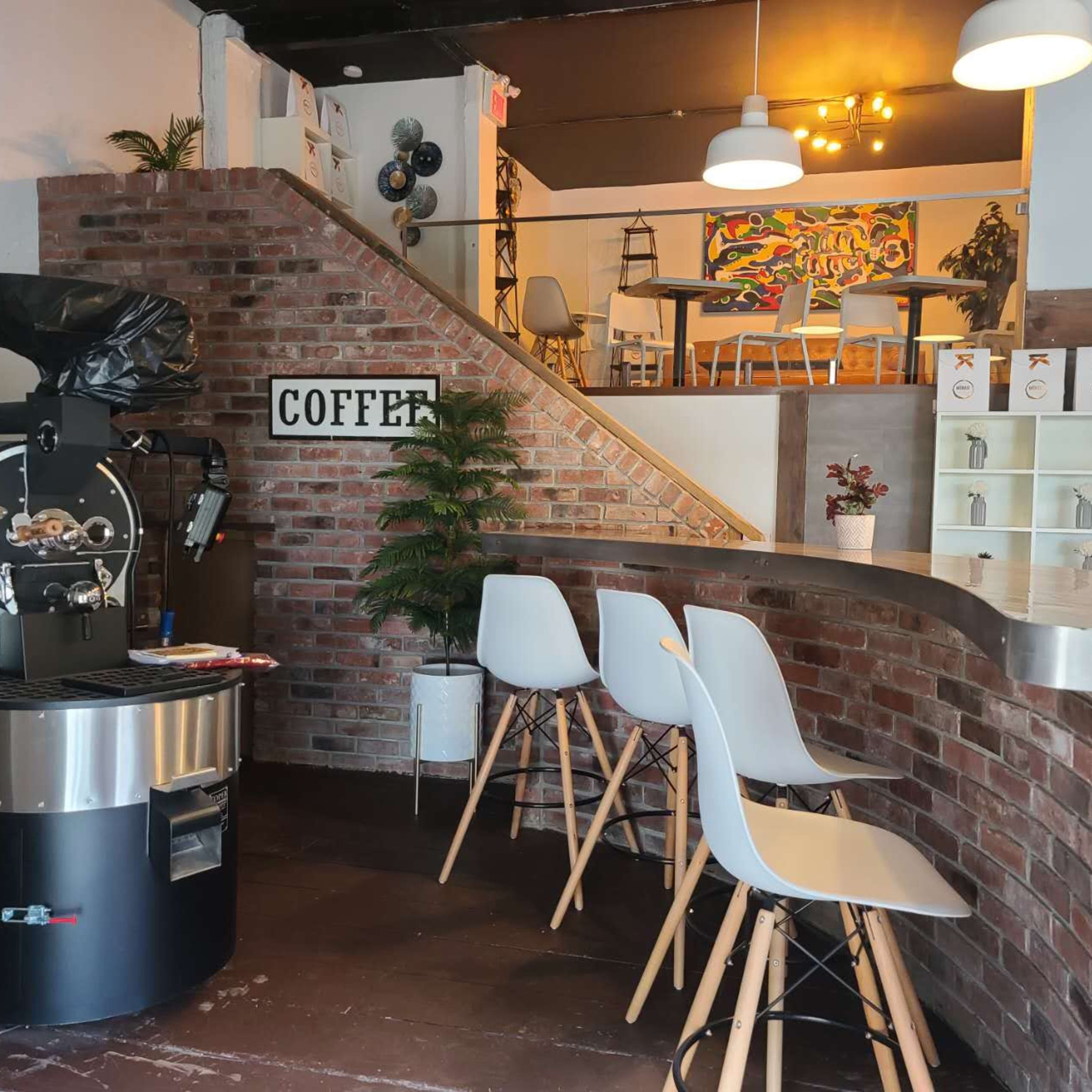 A coffee shop with a bar area featuring three white stools, a coffee roasting machine, and a staircase leading to an upper seating area.