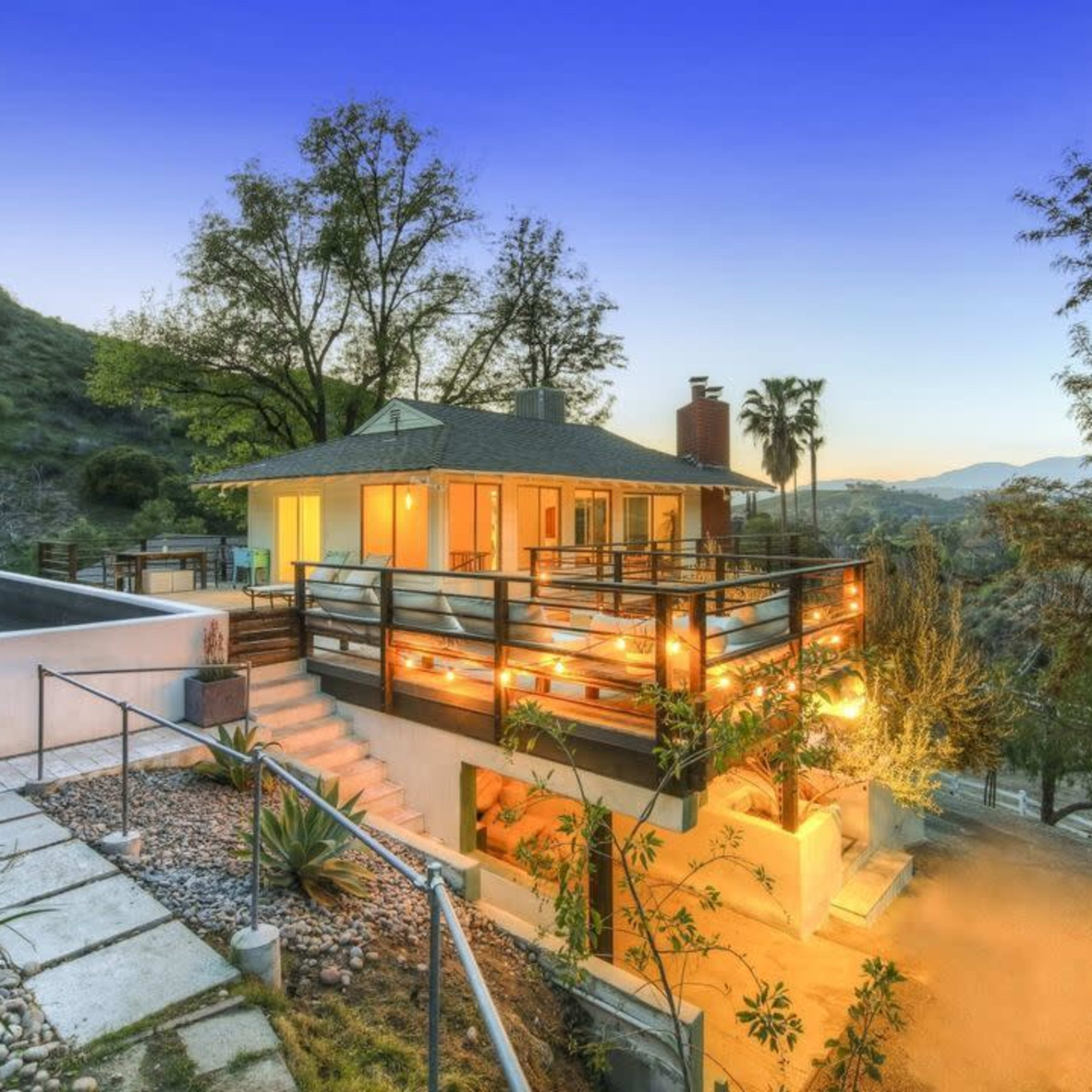 A modern house situated on a hillside, surrounded by greenery and visible outdoor lighting, with a pathway leading to the entrance.