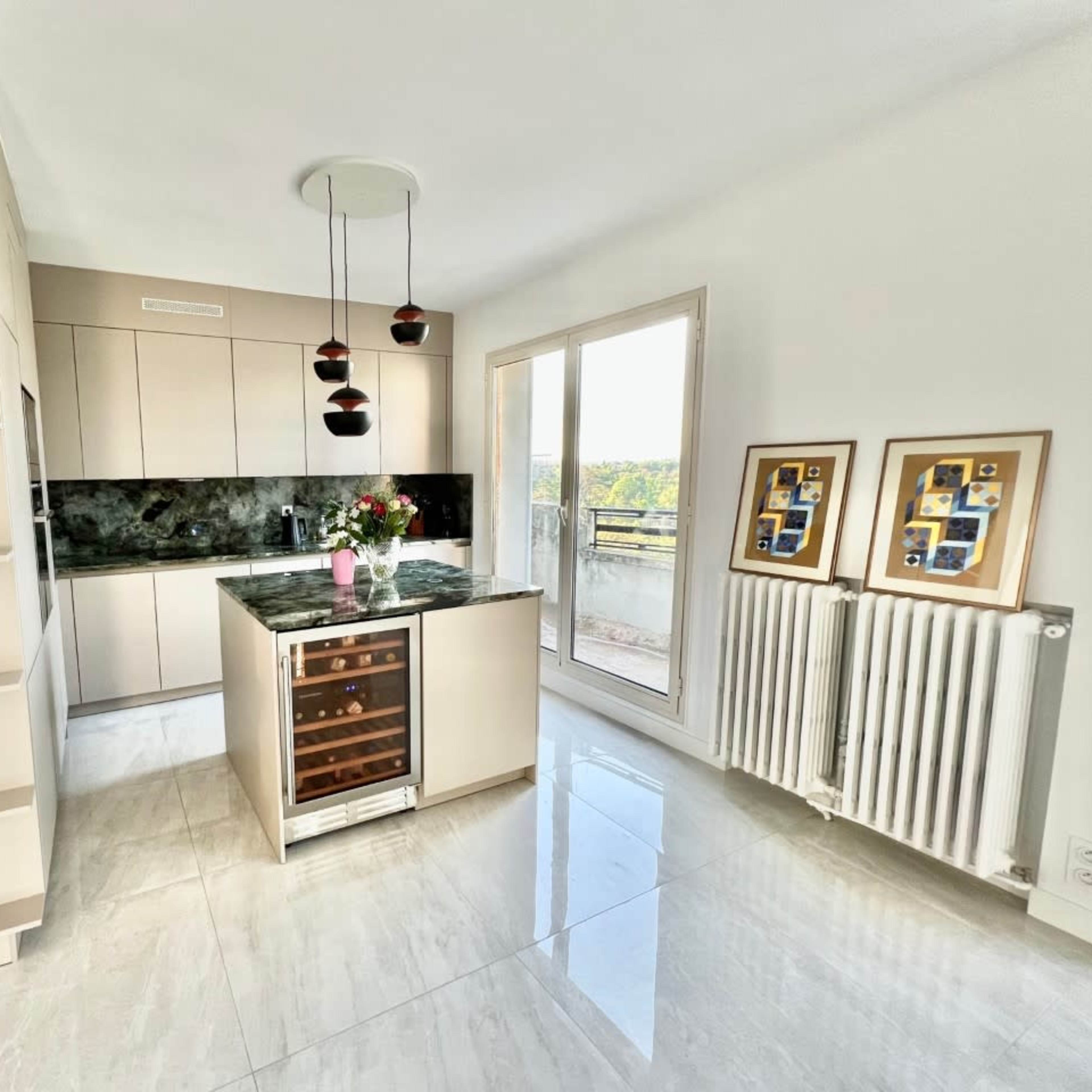 A modern kitchen with beige cabinets, a dark countertop, a wine cooler, and large windows that provide natural light.