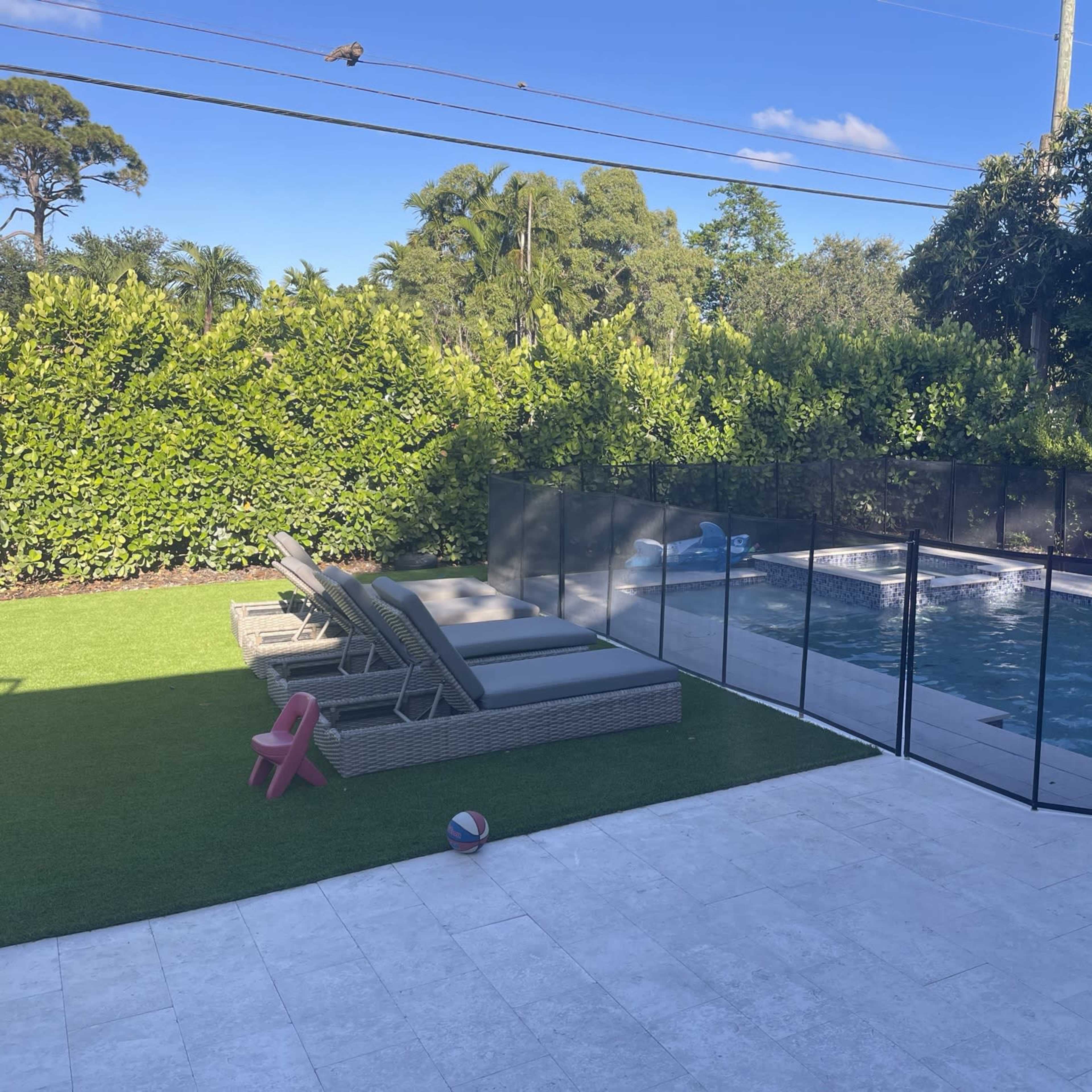 A backyard with a pool surrounded by a safety fence, lounge chairs, and a grassy area.