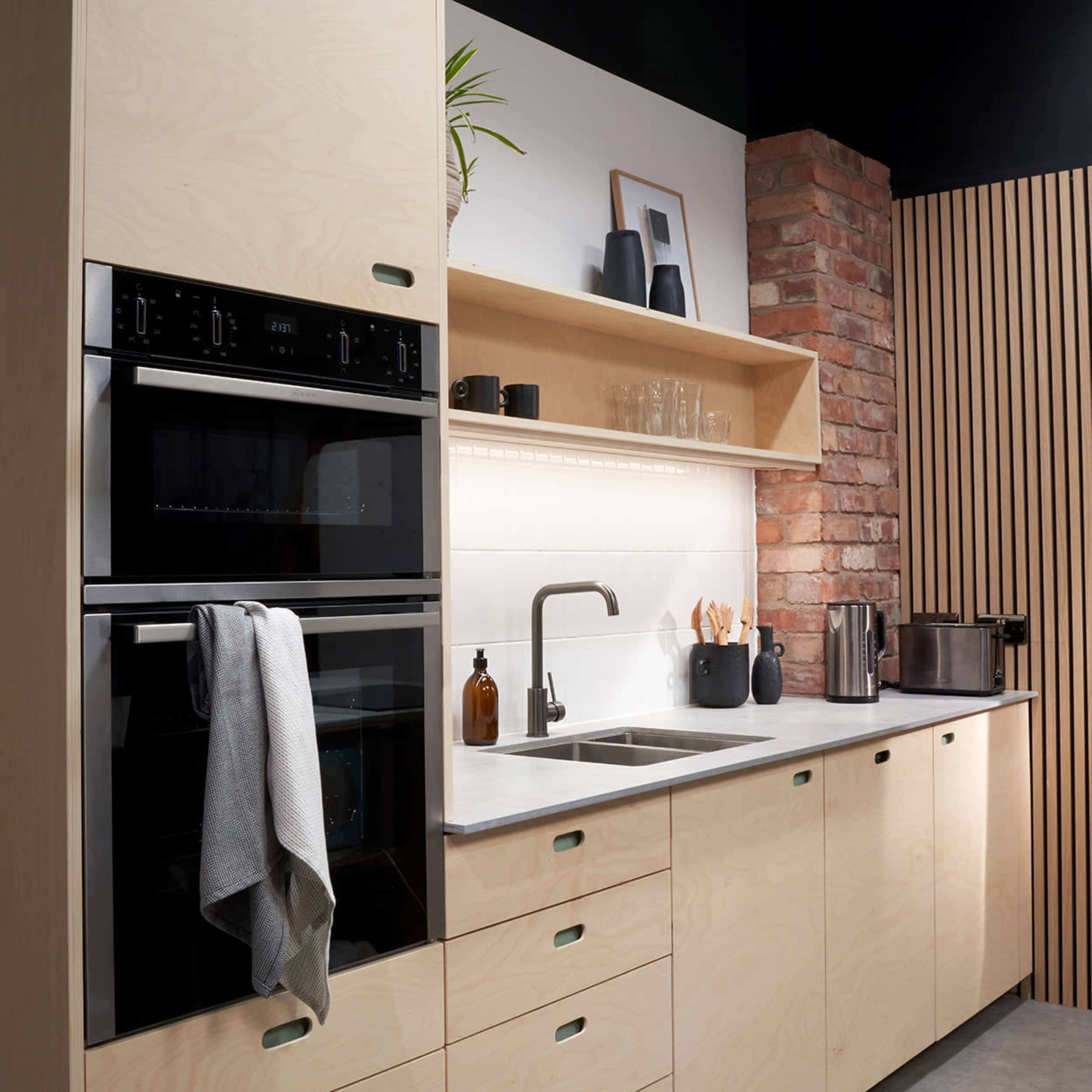 A modern kitchen featuring beige cabinetry, built-in black ovens, a minimalist sink, and a wooden accent wall.